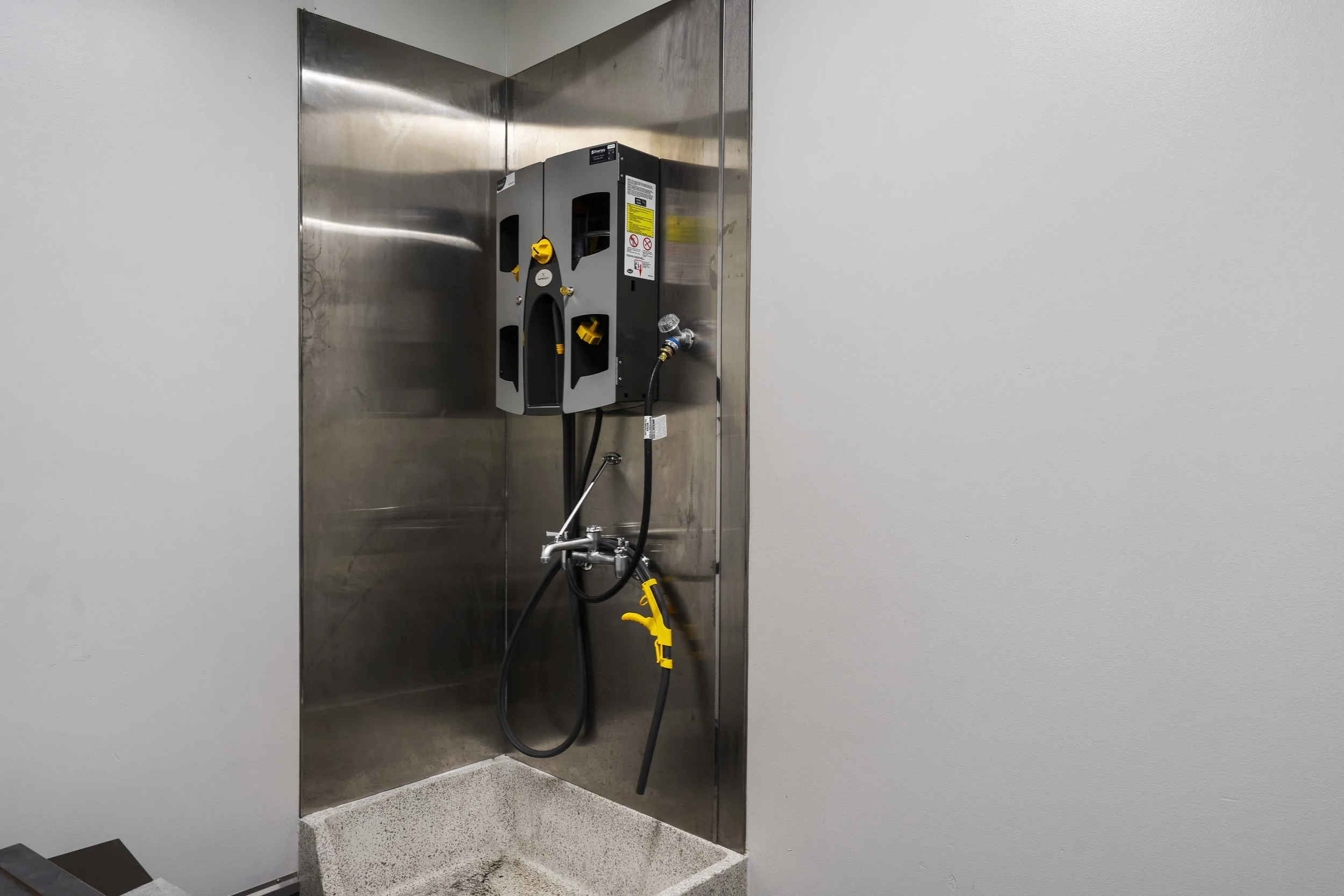 A wall-mounted electrical panel with safety warning labels, connected to a hose and spray nozzle, in a clean utility room with gray walls and a concrete floor.