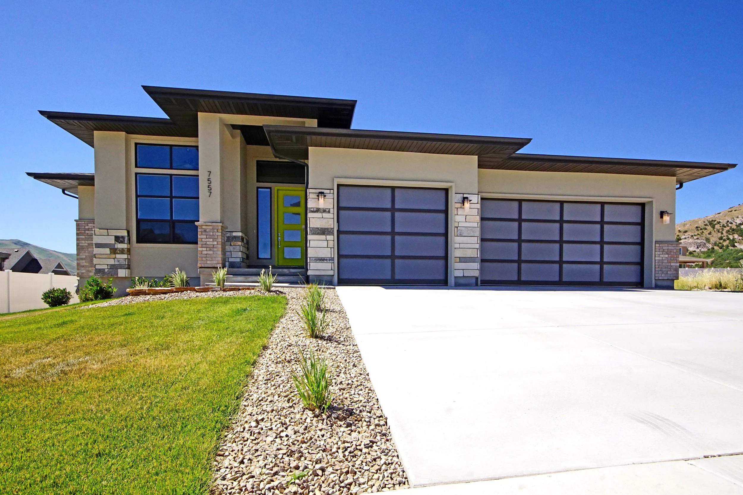 Modern single-story house with a wide driveway, large garage doors, and a distinctive green front door, surrounded by a well-maintained lawn and stone landscaping.