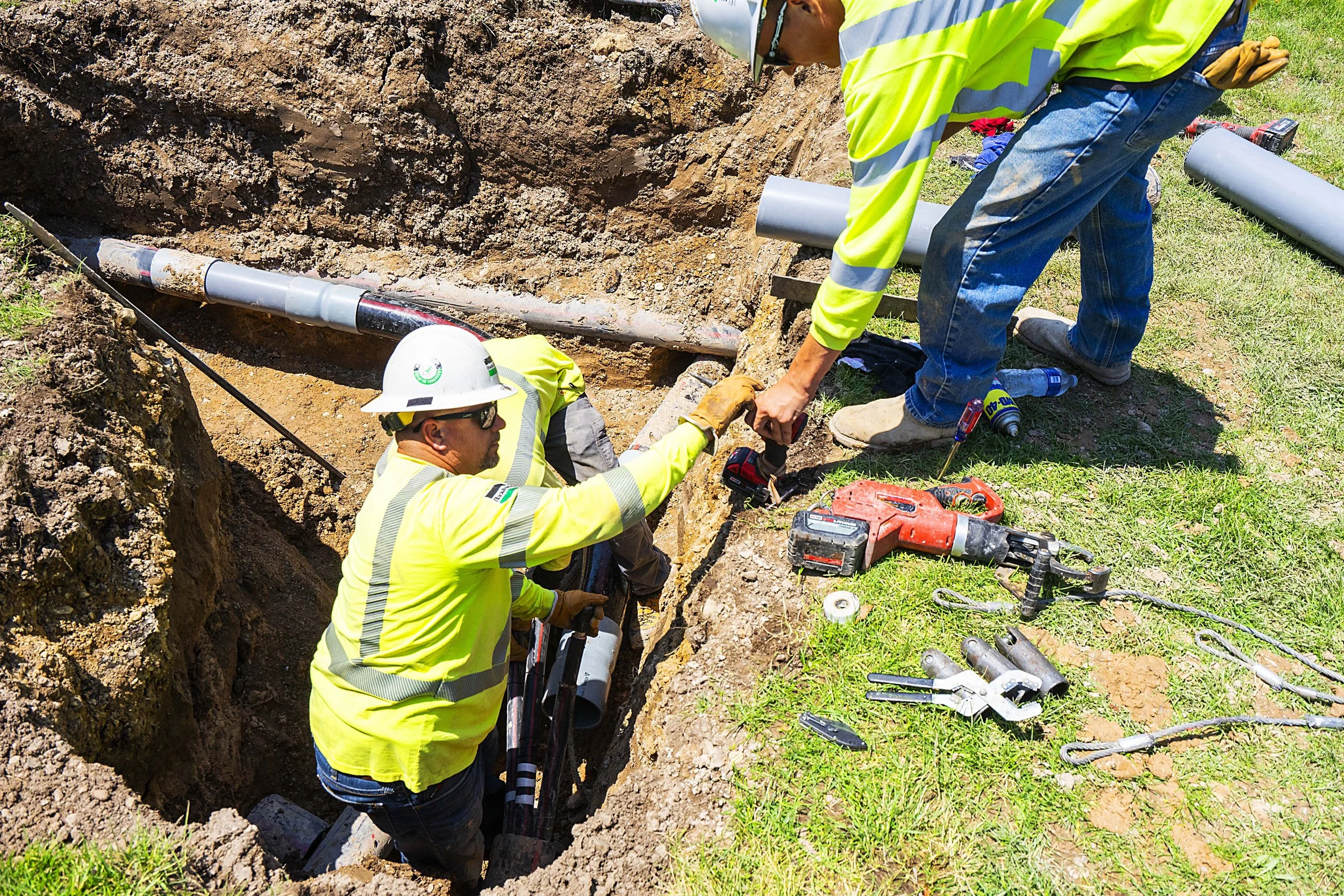 Construction workers installing underground pipes in a trench with various tools and equipment around them.