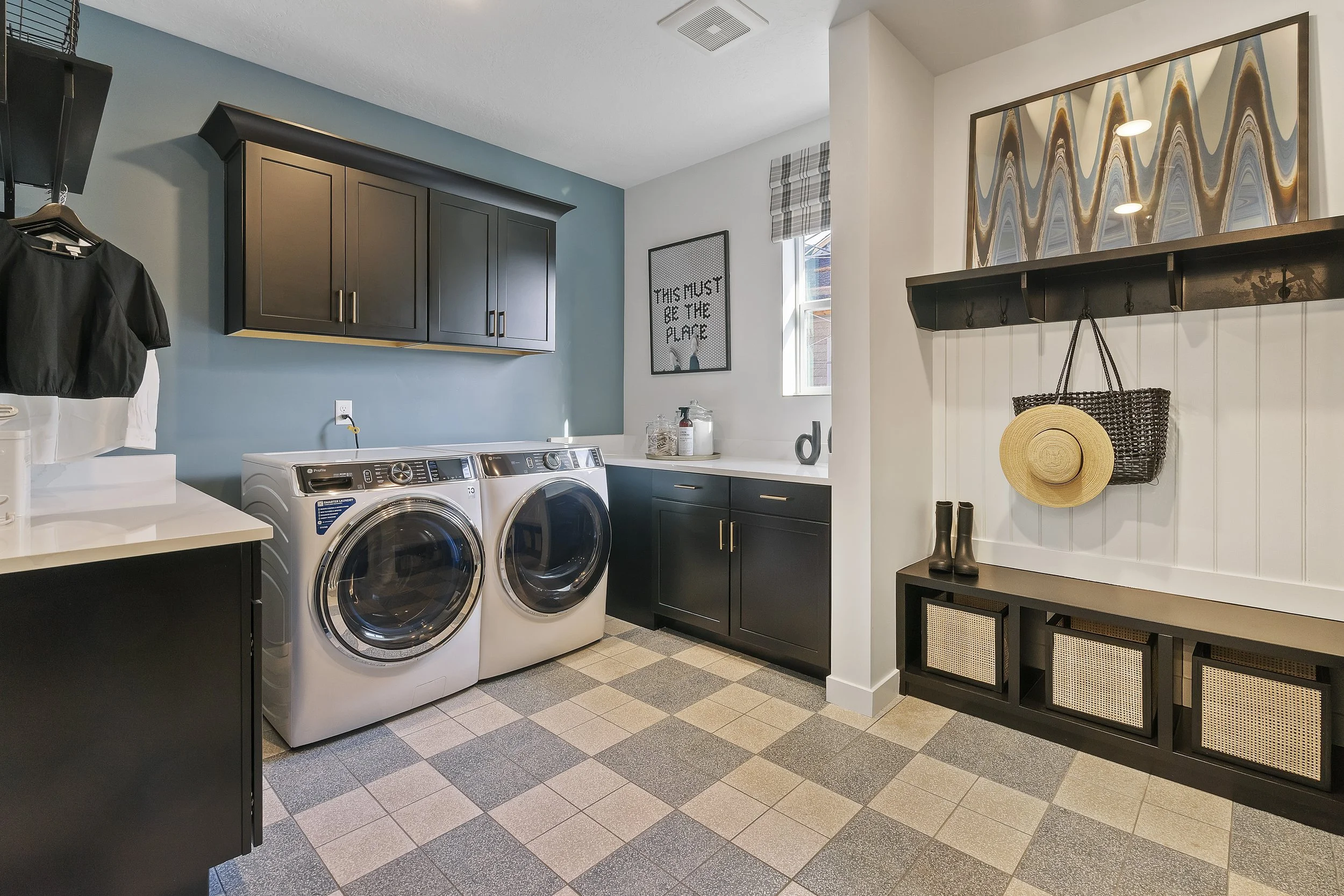 Laundry room with front-loading washer and dryer, black cabinets, a small counter, a window, framed artwork, and a black bench with woven baskets, boots, a hat, and hooks.