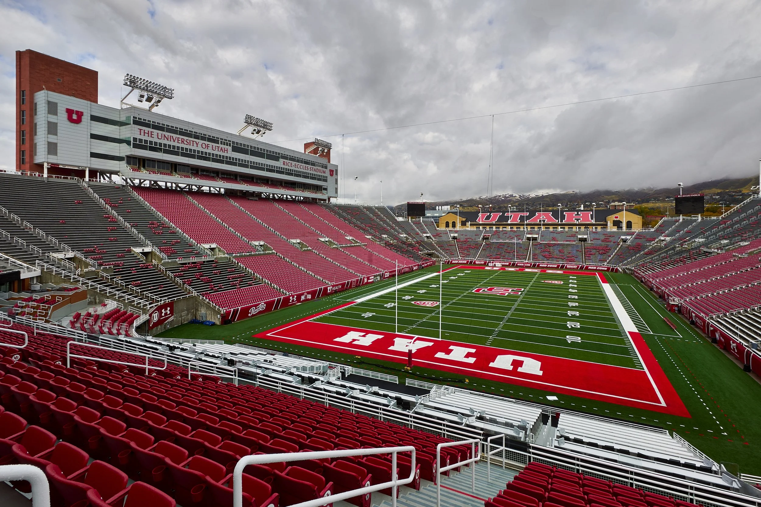 Empty football stadium at the University of Utah, with red seats, a green field, and a cloudy sky.