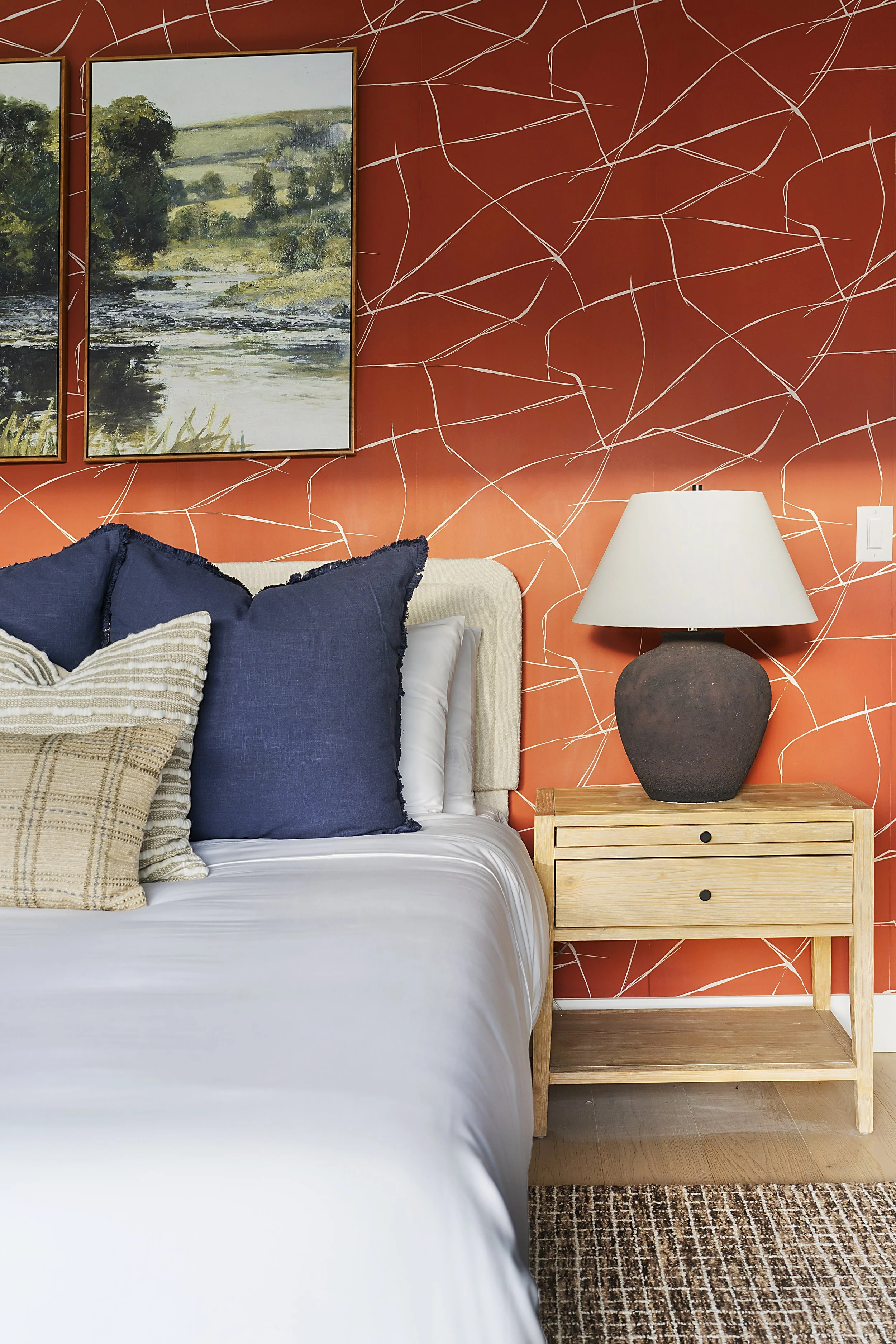 A bedroom with a beige headboard, navy and beige pillows on a white bed, an orange wall with white abstract lines, a landscape painting, a wooden nightstand, a dark ceramic table lamp with a white shade, and a brown and beige patterned rug.