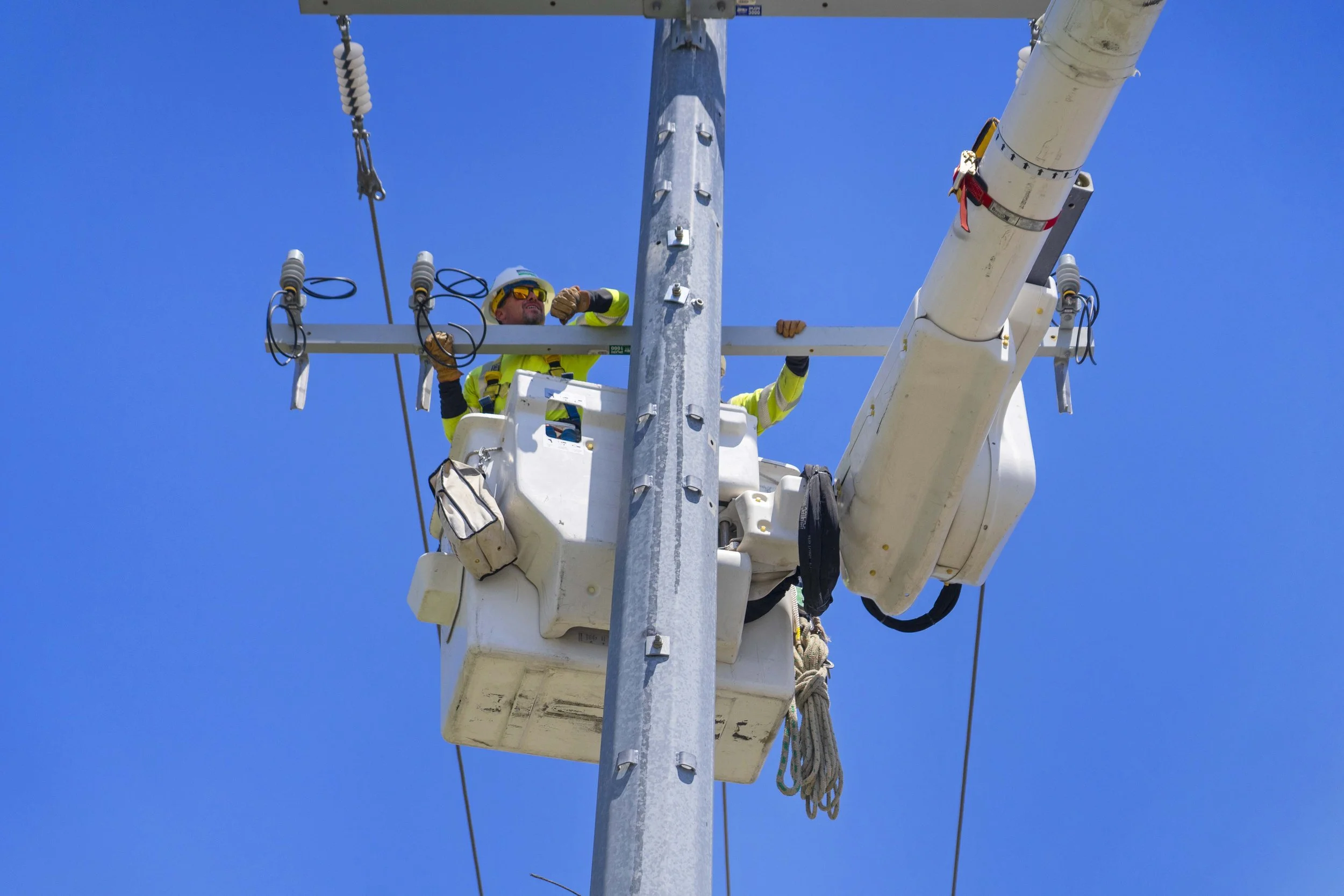 A worker wearing a helmet and reflective safety gear is in a bucket lift, working on a utility pole against a clear blue sky.