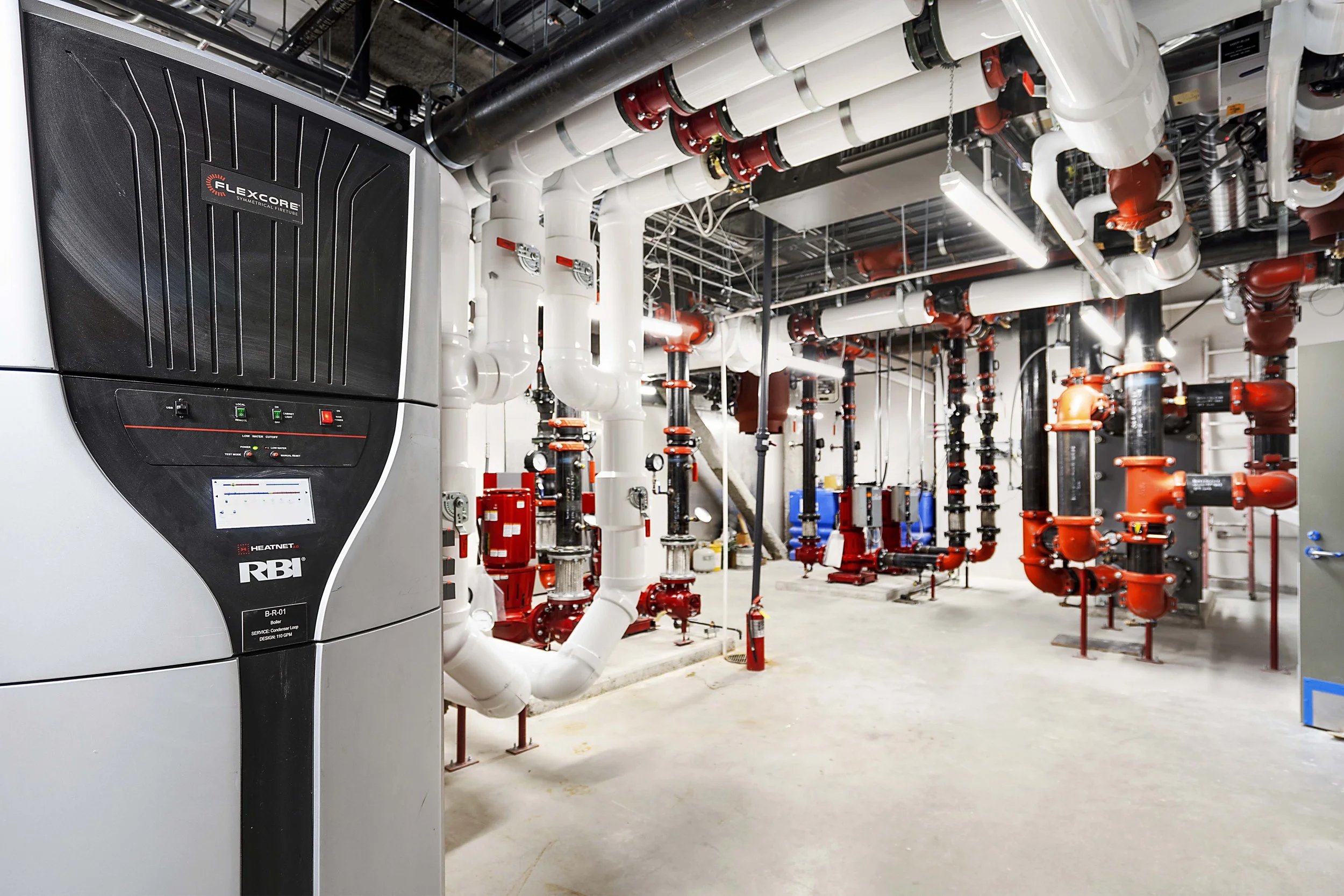 A mechanical room with an HVAC or boiler system, featuring white and red pipes, gauges, valves, and a large black and silver HVAC unit labeled 'Flexcore' and 'RBI'.