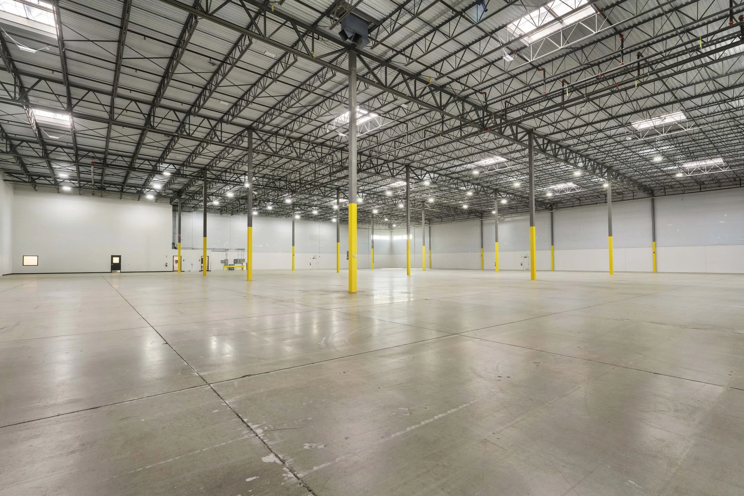 Empty warehouse with high ceiling, metal framework, concrete floor, and yellow safety poles.