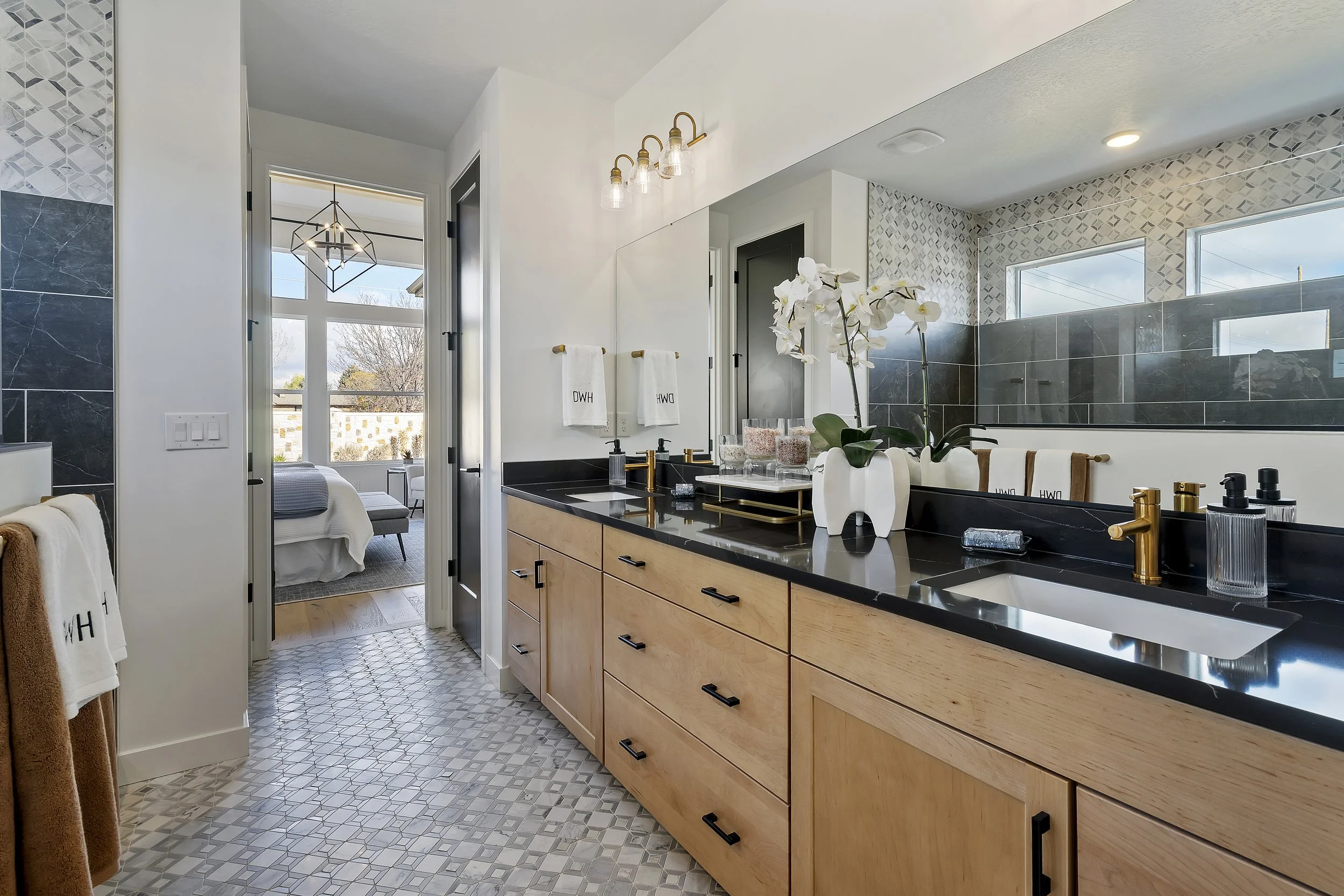 Modern bathroom with wooden vanity, black countertop, white sink, gold fixtures, decorative orchids, and a large mirror. Through the open door, a bedroom with a bed, sitting area, and large window is visible.