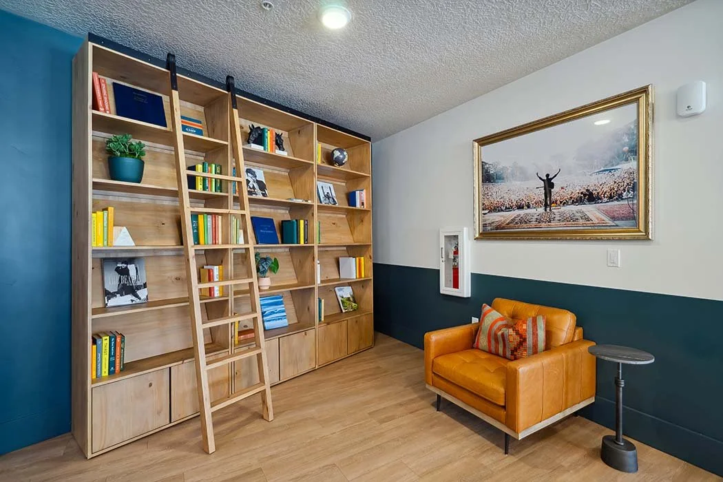 Living room with a tall wooden bookshelf, ladder, orange armchair with pillows, framed artwork of a person with arms raised, and a small side table.