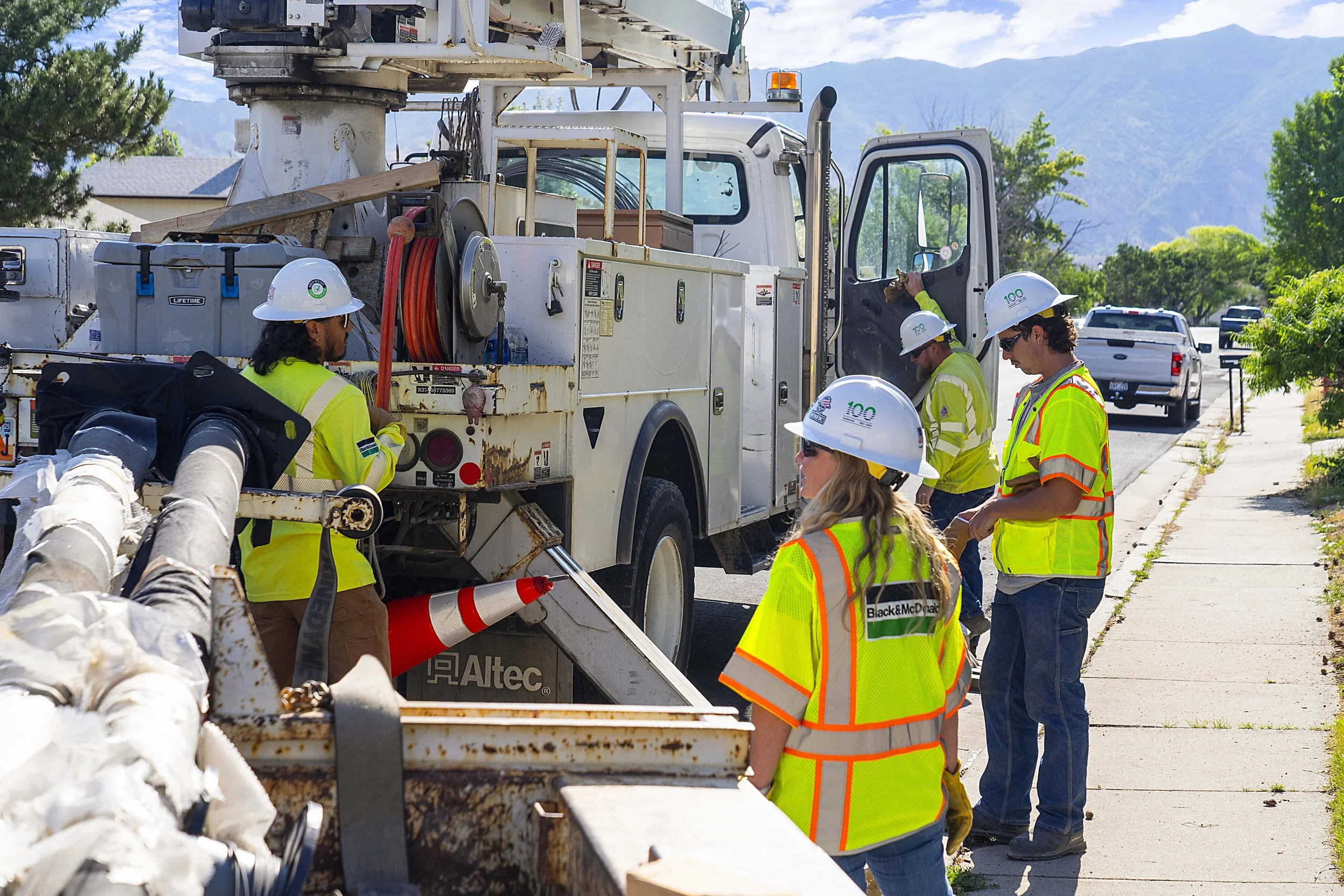 Group of utility workers wearing safety vests and helmets working outdoors on a white utility truck, with a suburban neighborhood and mountains in the background.