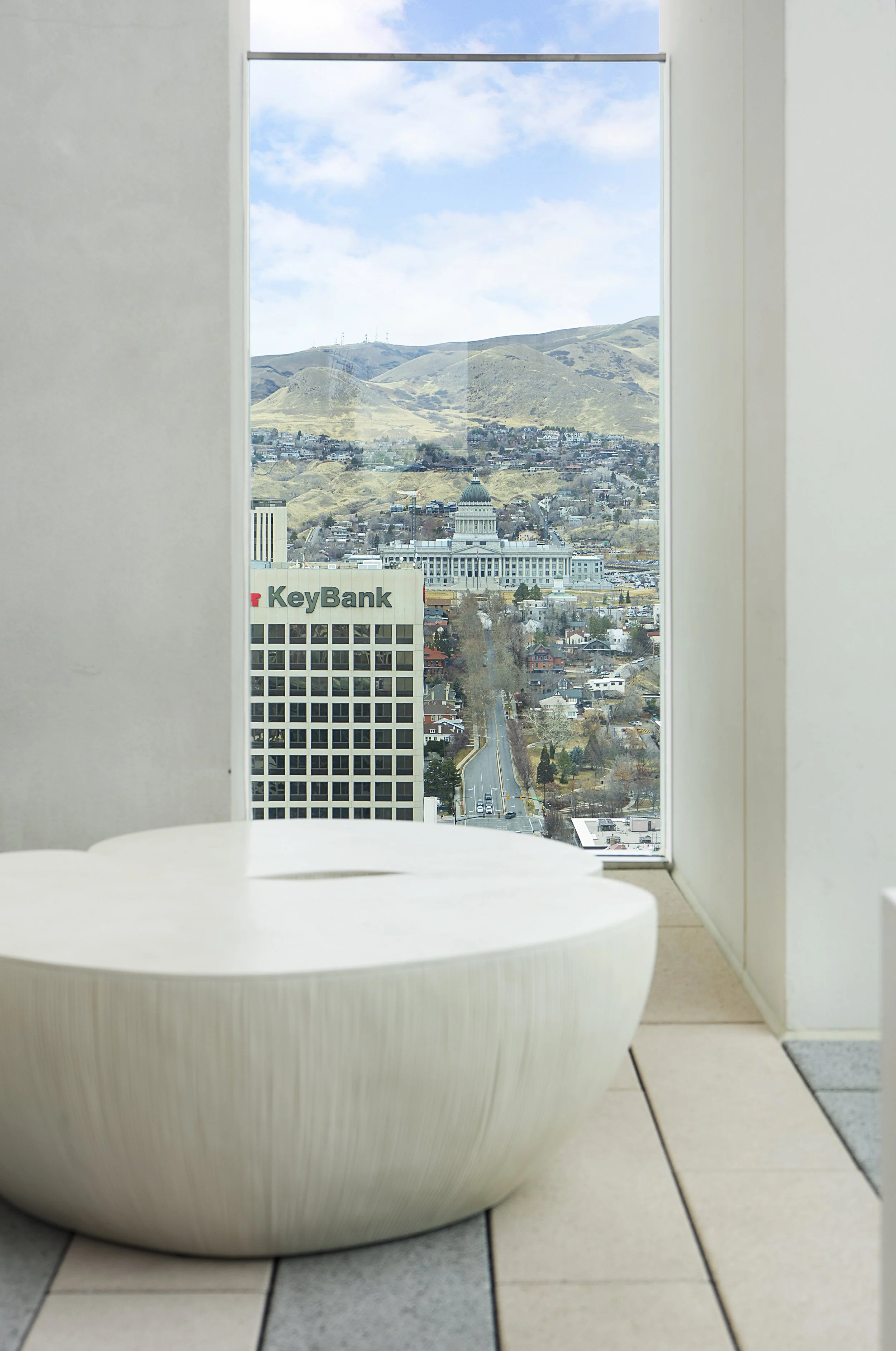 Interior of a building with a round white seating area and a large window overlooking a cityscape and distant hills.