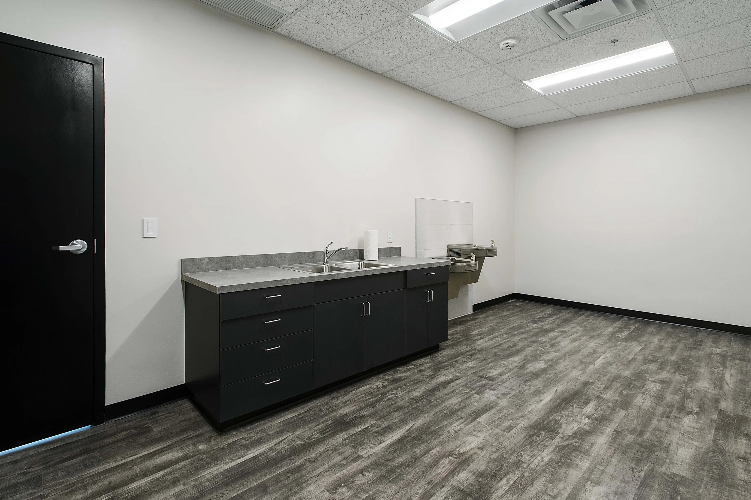 Empty kitchenette area with black cabinets, a sink, paper towel roll, and a water fountain in a room with white walls, gray flooring, and ceiling lights.