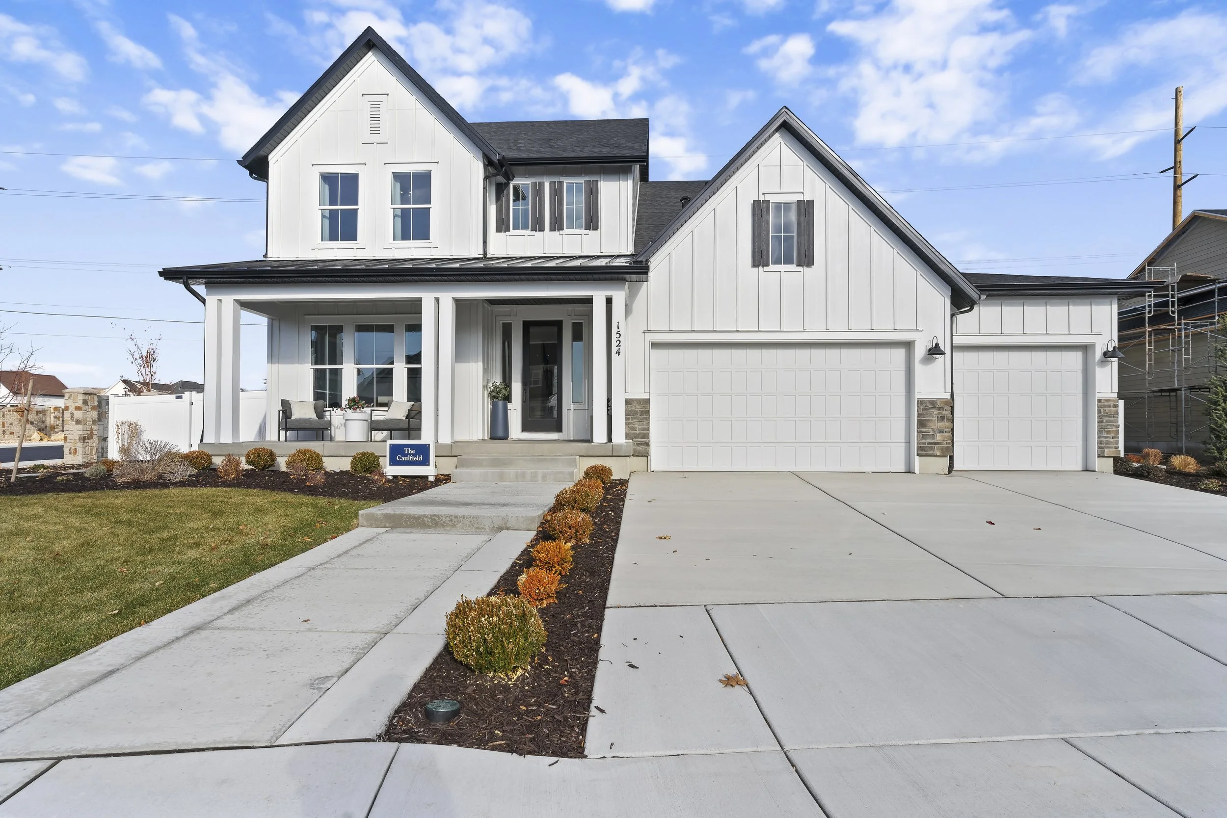 Modern two-story white house with black roof, front porch with seating area, dual garage doors, manicured lawn, walkway, and landscaping with small bushes.