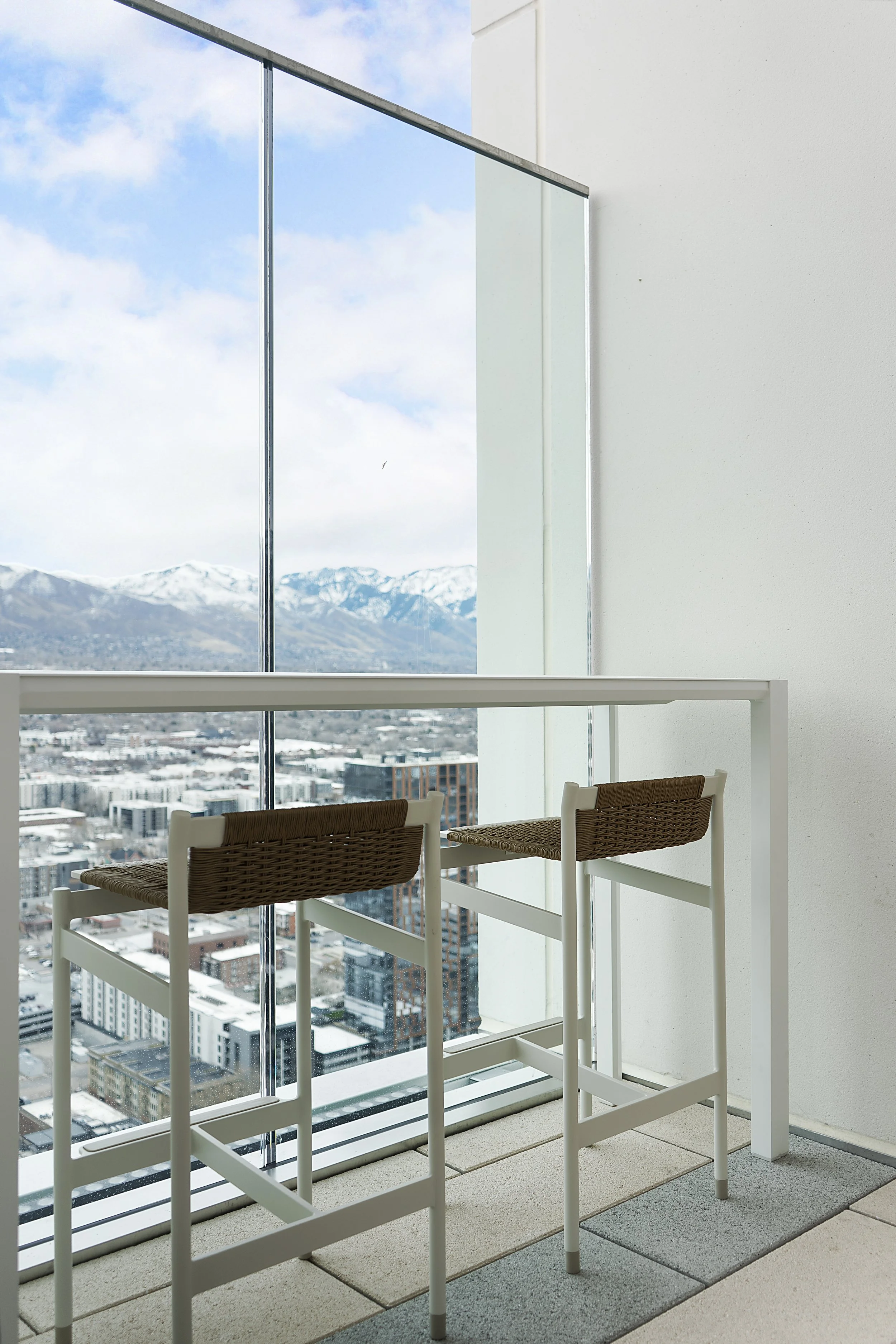A modern high-rise apartment balcony with two white bar stools with woven seats, overlooking a cityscape with snow-capped mountains in the background.