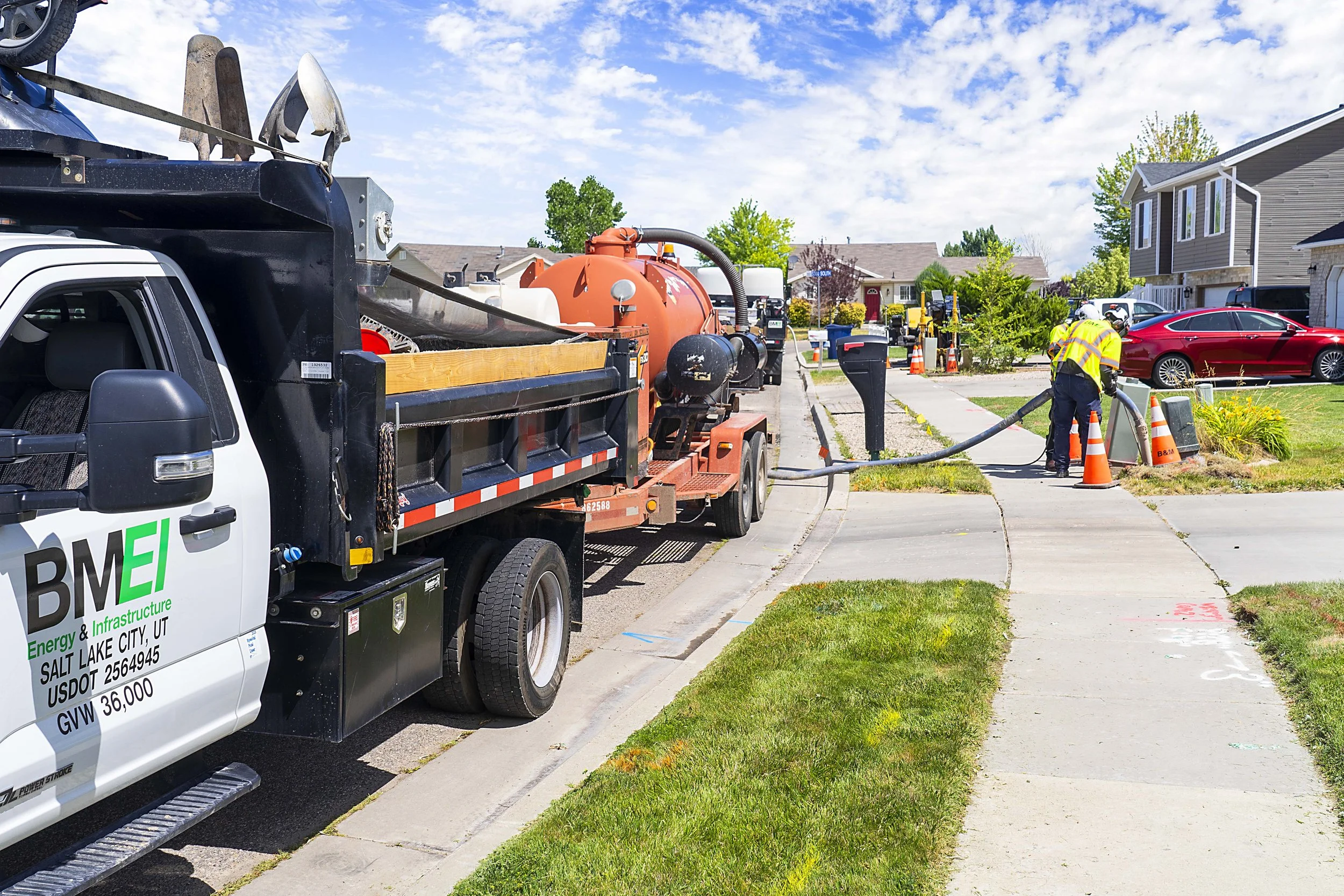 Workers in yellow safety vests manage a sewer or drain cleaning operation on a suburban sidewalk with a truck and a large hose.