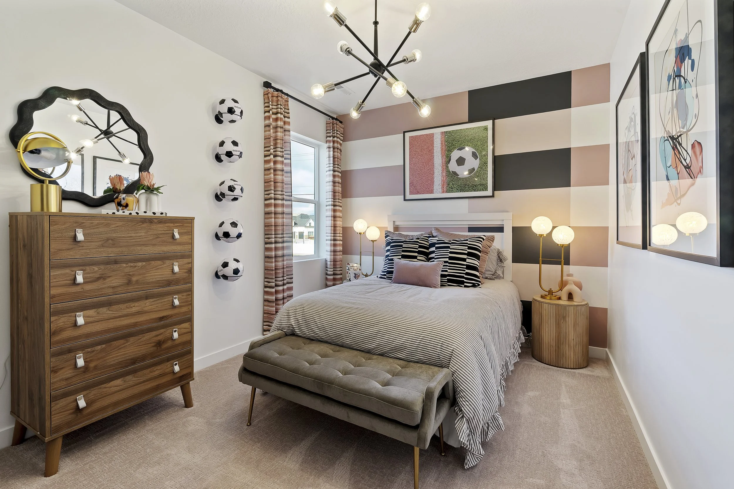 A bedroom with a football theme decor, featuring a bed with striped pillows, a striped throw blanket, and football wall art. The room has a striped accent wall, a wooden dresser with a mirror, and a large window with curtains. A bench is at the foot 