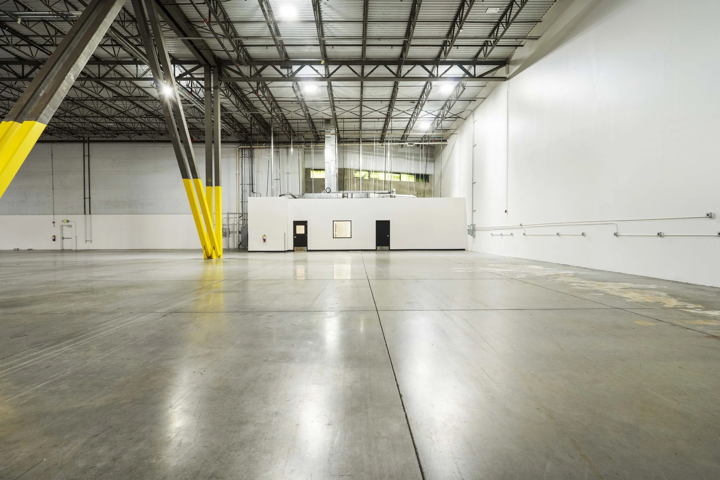 Empty warehouse with high ceiling, exposed steel beams, and polished concrete floor. There is a small enclosed white room with doors and windows at the back, and a yellow and black safety post supporting a large steel beam on the left side.