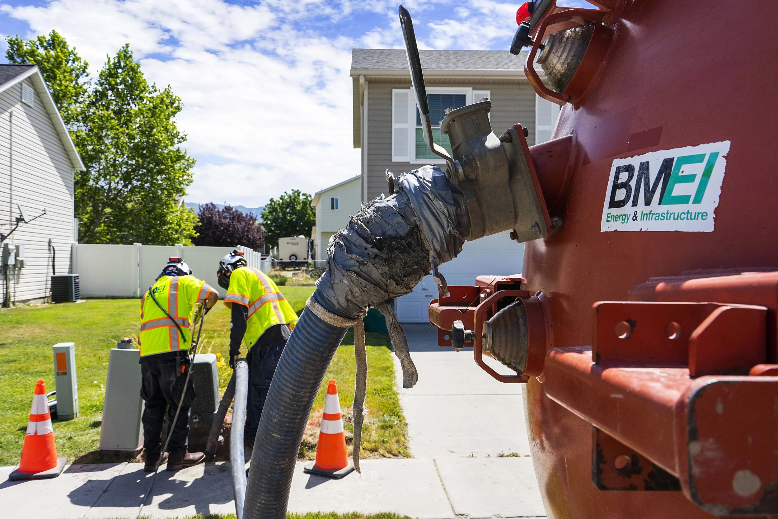 Three workers in yellow safety vests and helmets working outdoors with a large orange utility truck labeled BMEI, beside orange traffic cones and a residential house in the background.