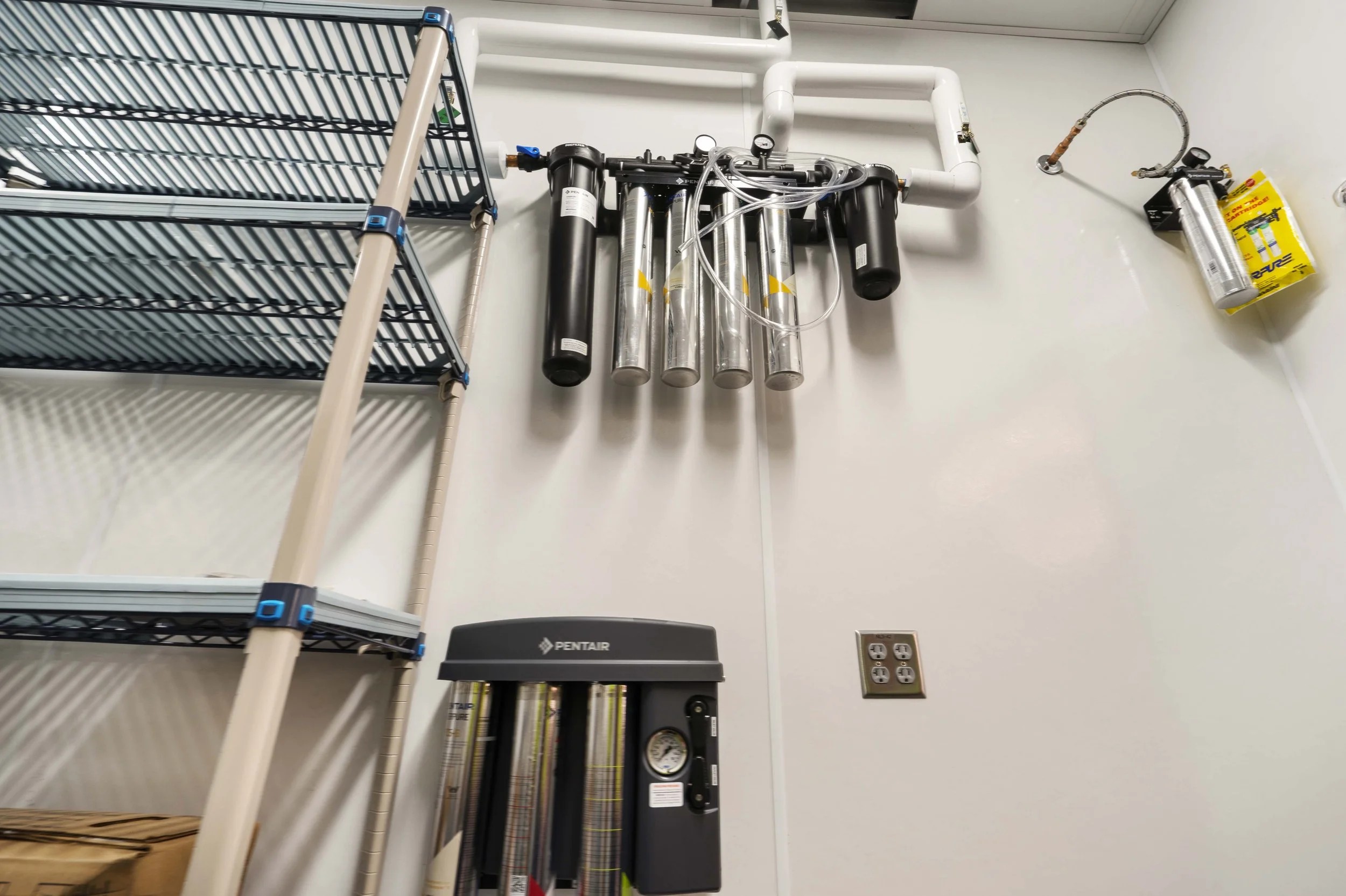 Medical or laboratory equipment mounted on a white wall, including water filtration or ozonation system, a fire extinguisher, and a metal shelving unit.