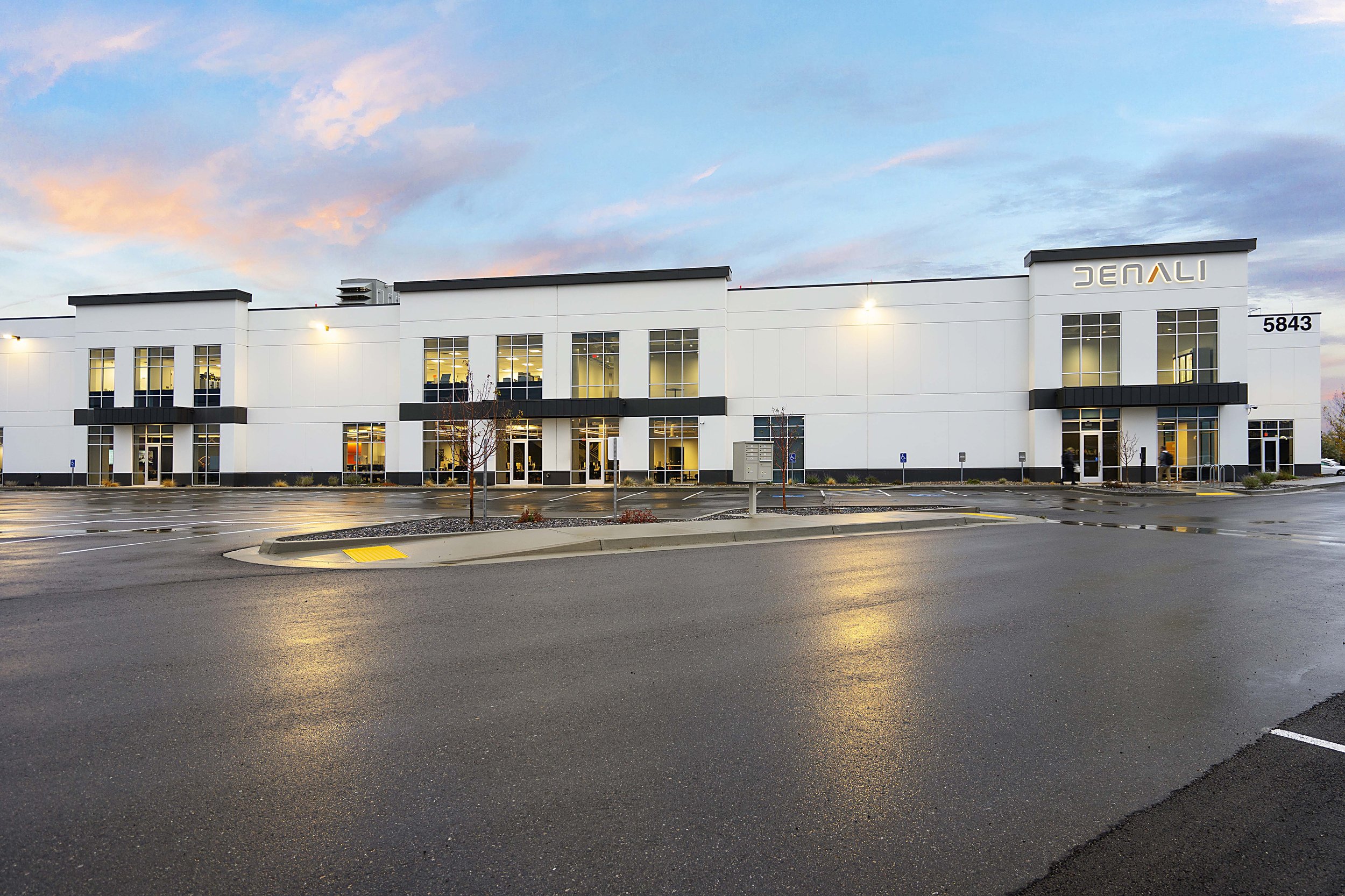 Modern commercial building with white exterior, large windows, and multiple entrance doors. Parking lot in front and a sky with soft pink and blue clouds.