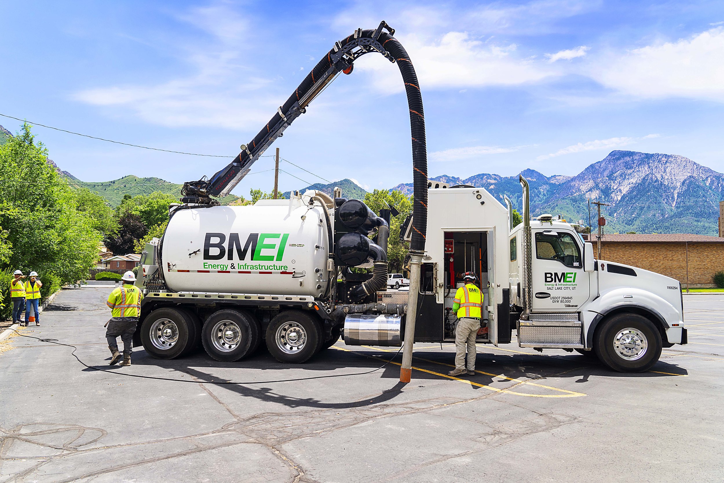 Workers in safety vests operating a large BME Energy & Infrastructure vacuum truck with a hose extended to a utility pole in a parking lot against a mountainous background.