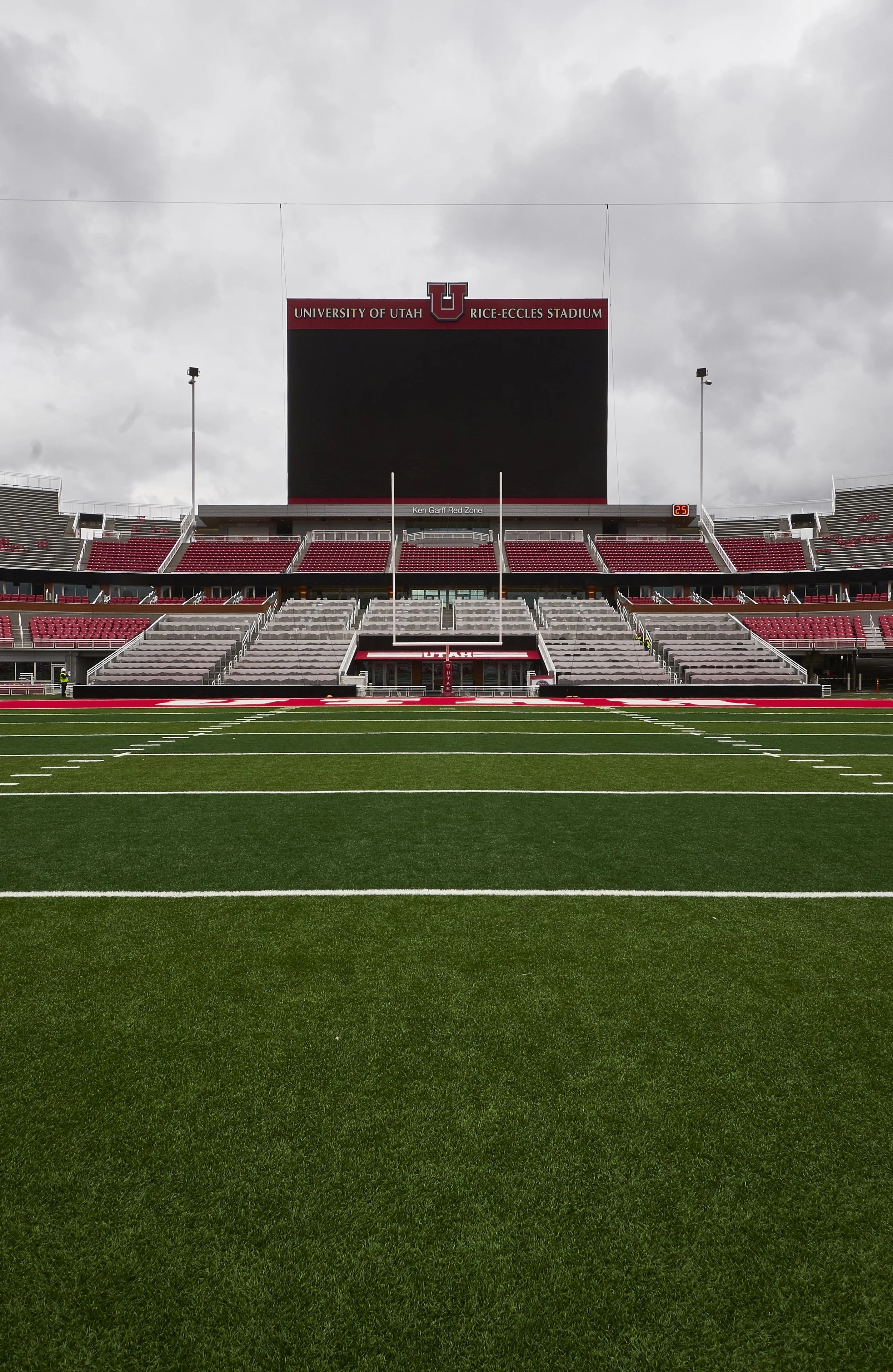 A view of Rice-Eccles Stadium at the University of Utah, with green football field in the foreground, empty red and gray bleachers, and a large scoreboard under a cloudy sky.