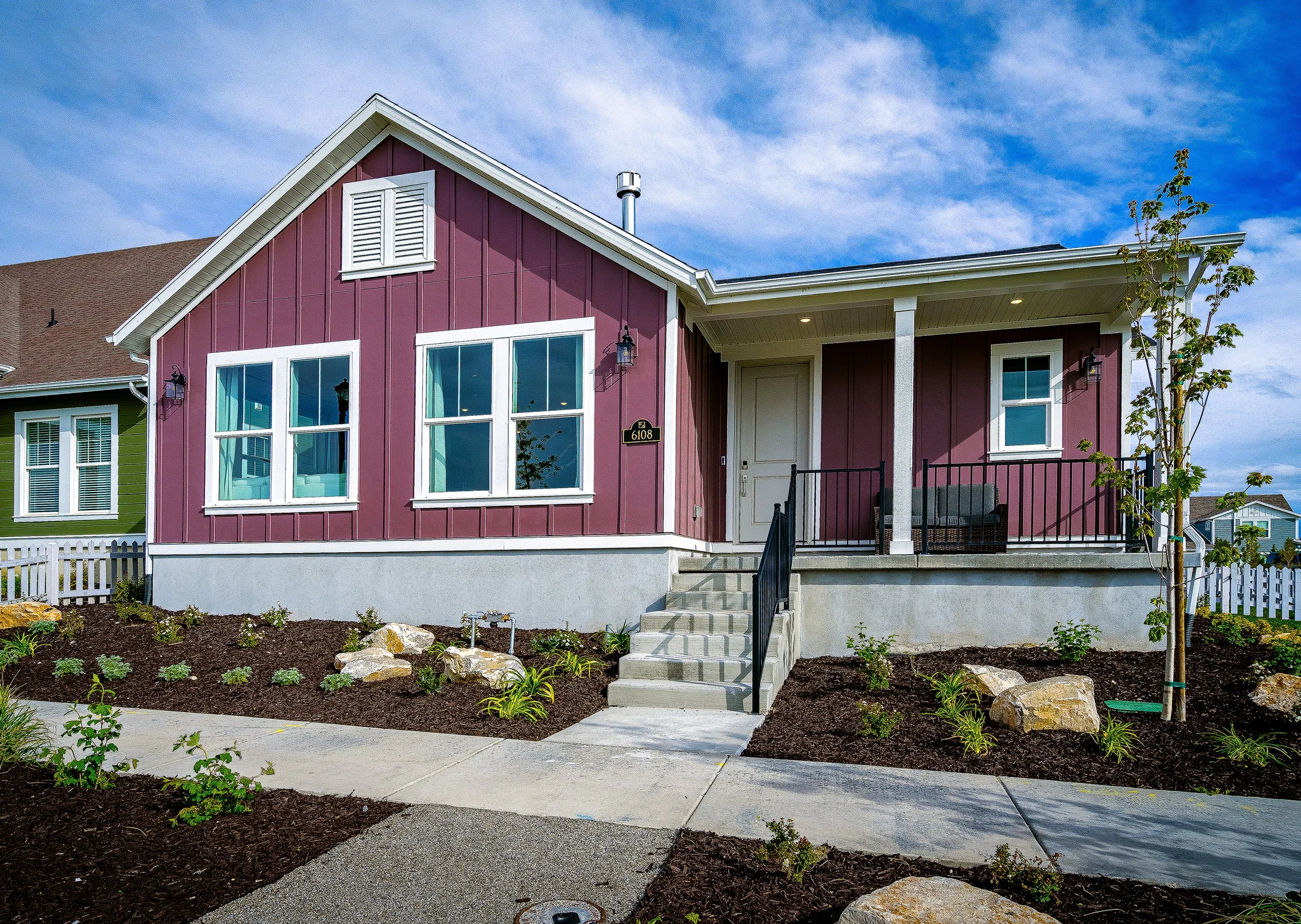 Single-story house with a red exterior, white trim, and covered porch. Fronted by a landscaped yard with small plants and rocks, under a blue sky with clouds.