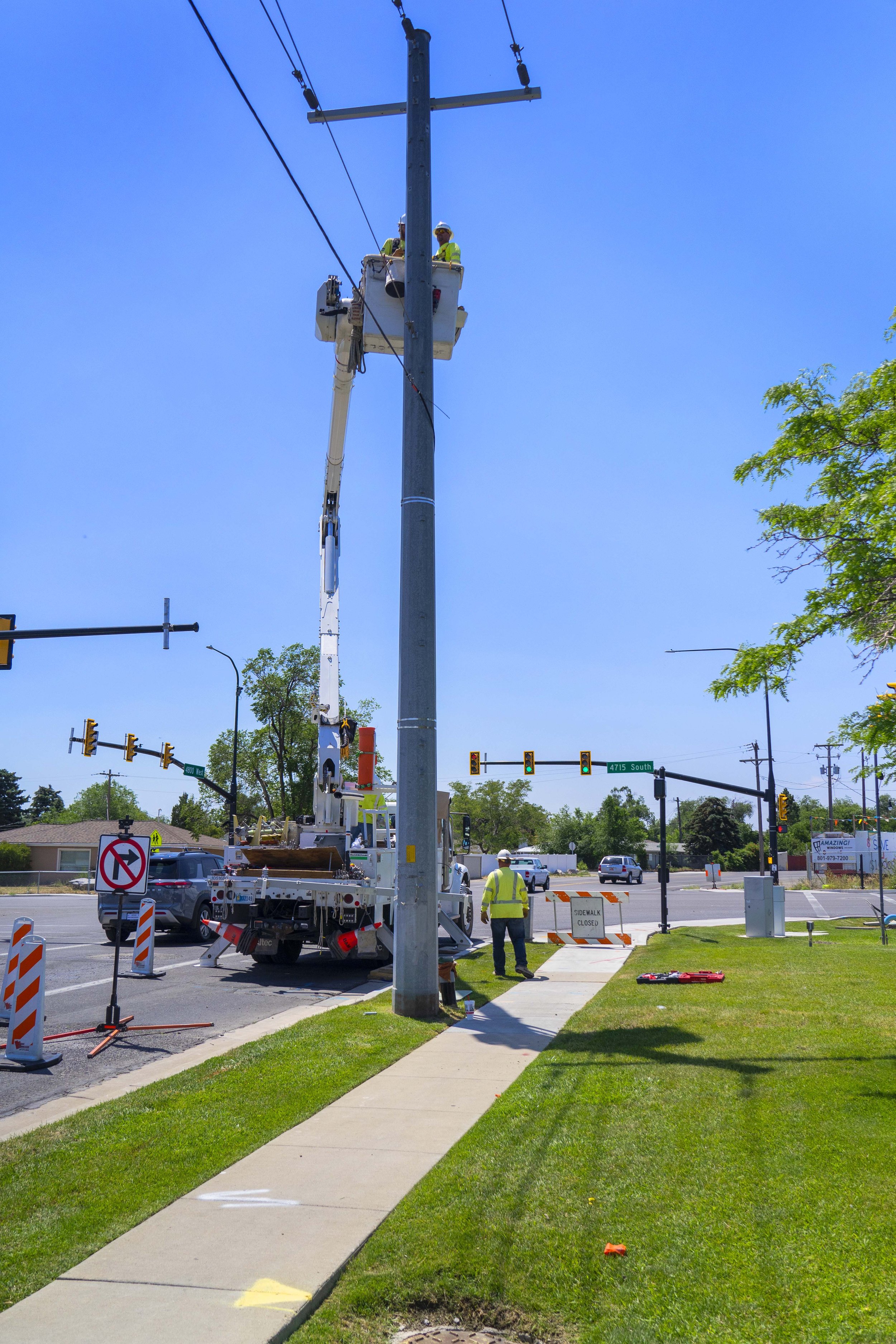 Workers repairing or maintaining a utility pole using a truck with a lift at a street corner under a clear blue sky.