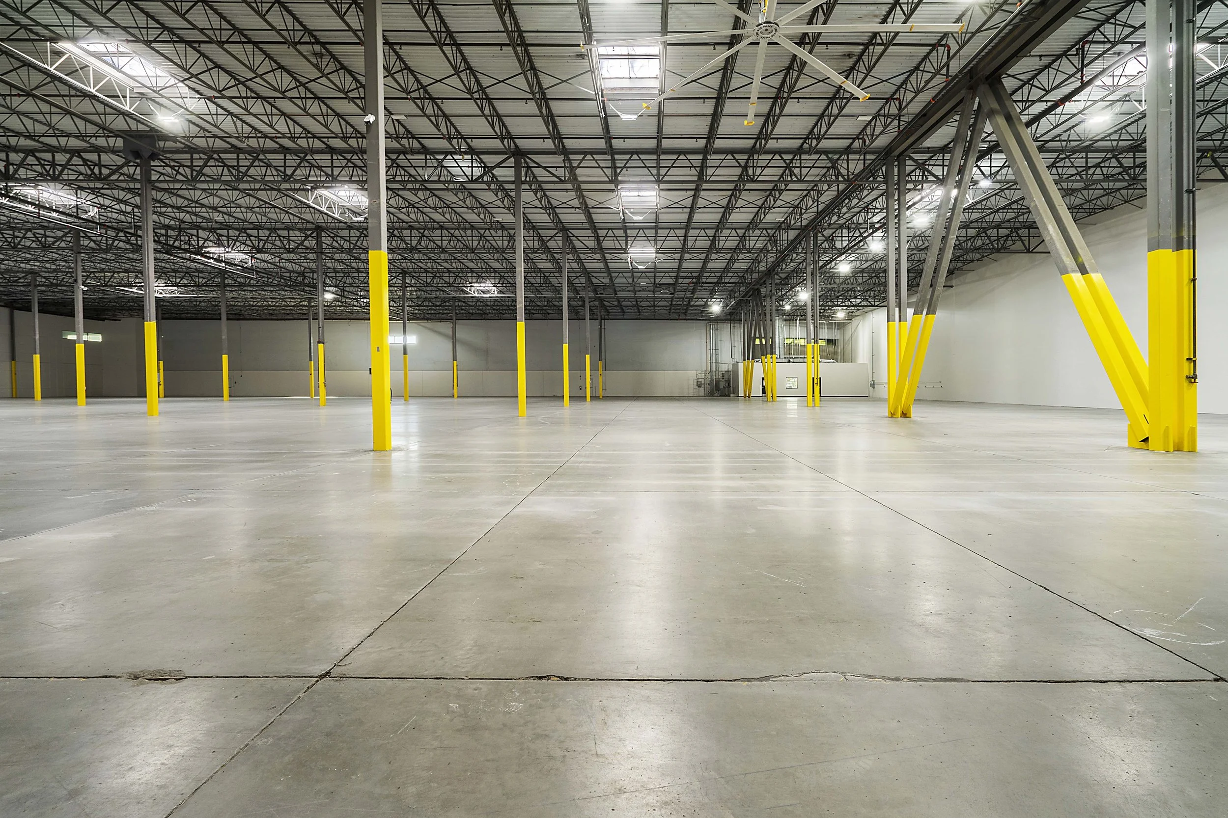 Empty warehouse with concrete floor, yellow safety posts, and metal roof structure with lighting.