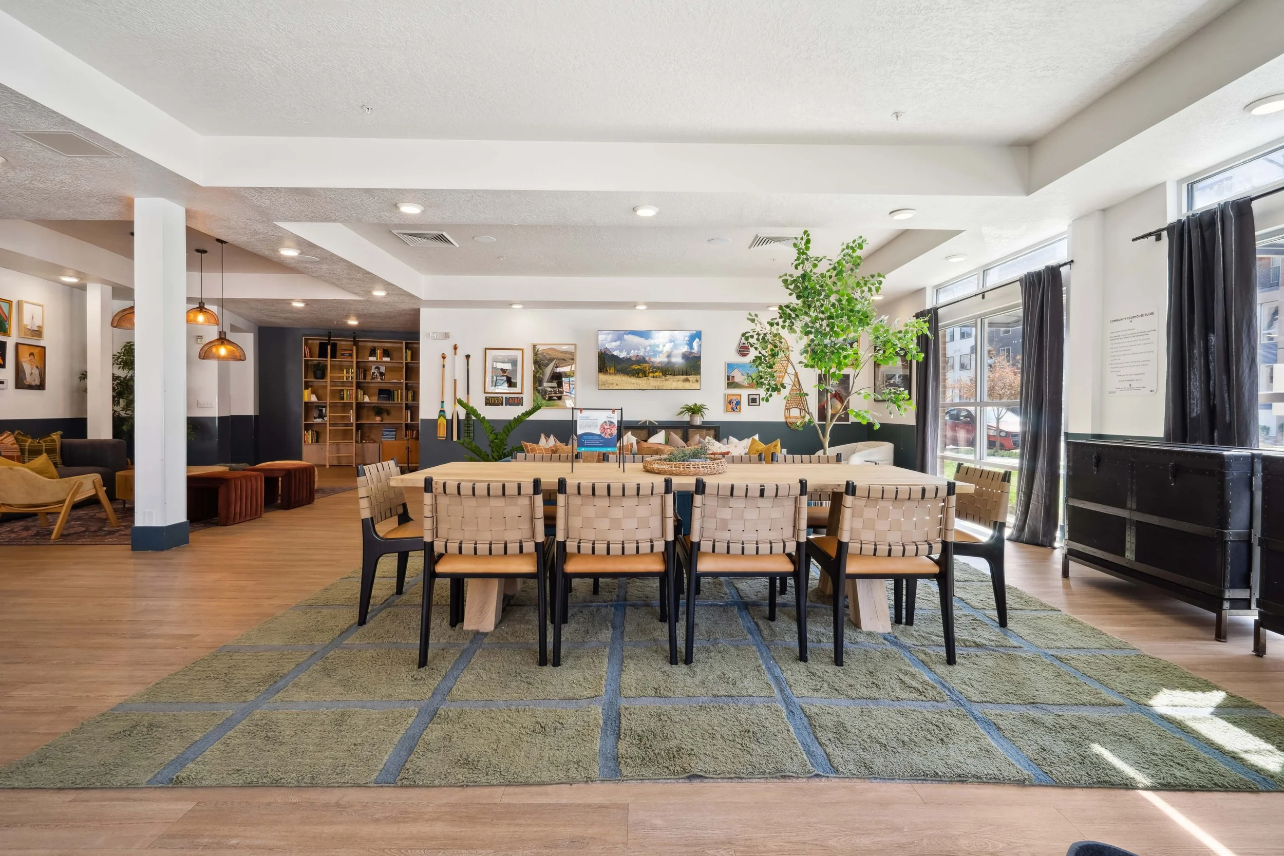 Bright and spacious indoor communal area with a large wooden dining table surrounded by woven and black chairs, a green rug underneath, and large windows with black curtains on the right. Decor includes potted plants, wall art, and a bookshelf in the