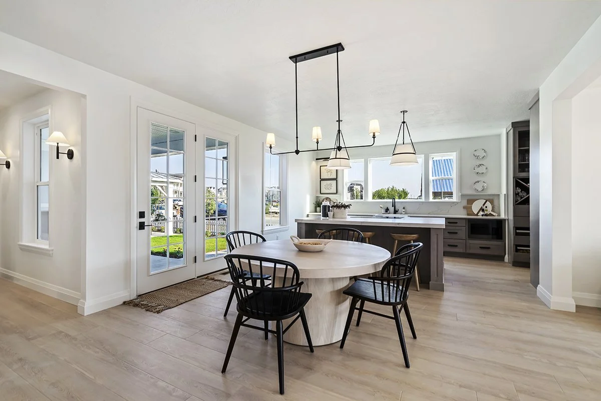 Bright, open-concept kitchen and dining area with white walls, light wood floors, black chairs, a round white dining table, and modern light fixtures, with large windows and French doors bringing in natural light.