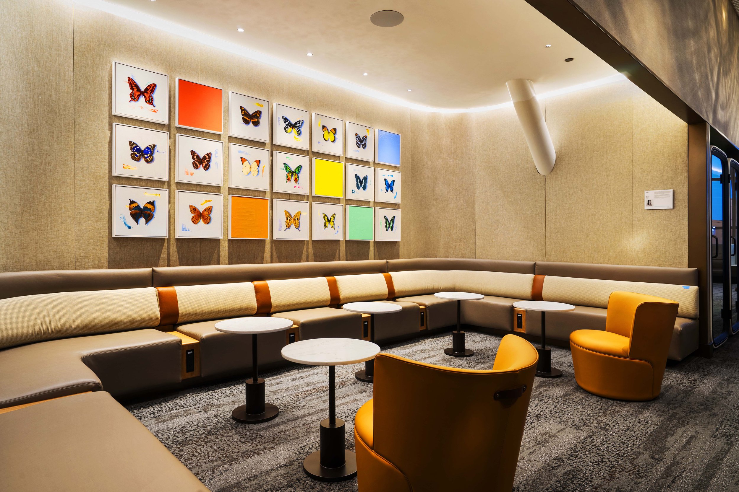Modern lounge area with beige sectional sofa, orange armchairs, and small round tables. Art display of colorful butterfly illustrations on the wall.