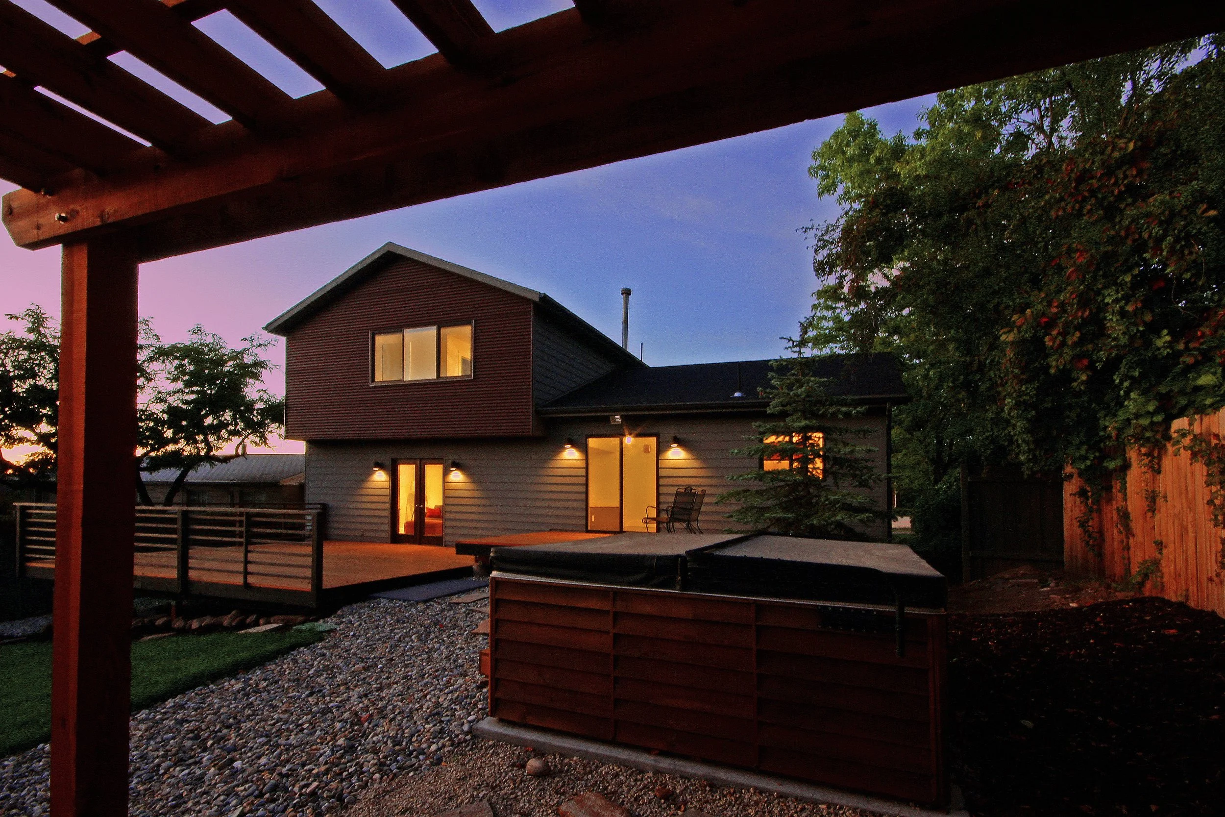 Modern house exterior with deck at dusk, surrounded by trees and a wooden fence, with patio furniture visible on the deck.