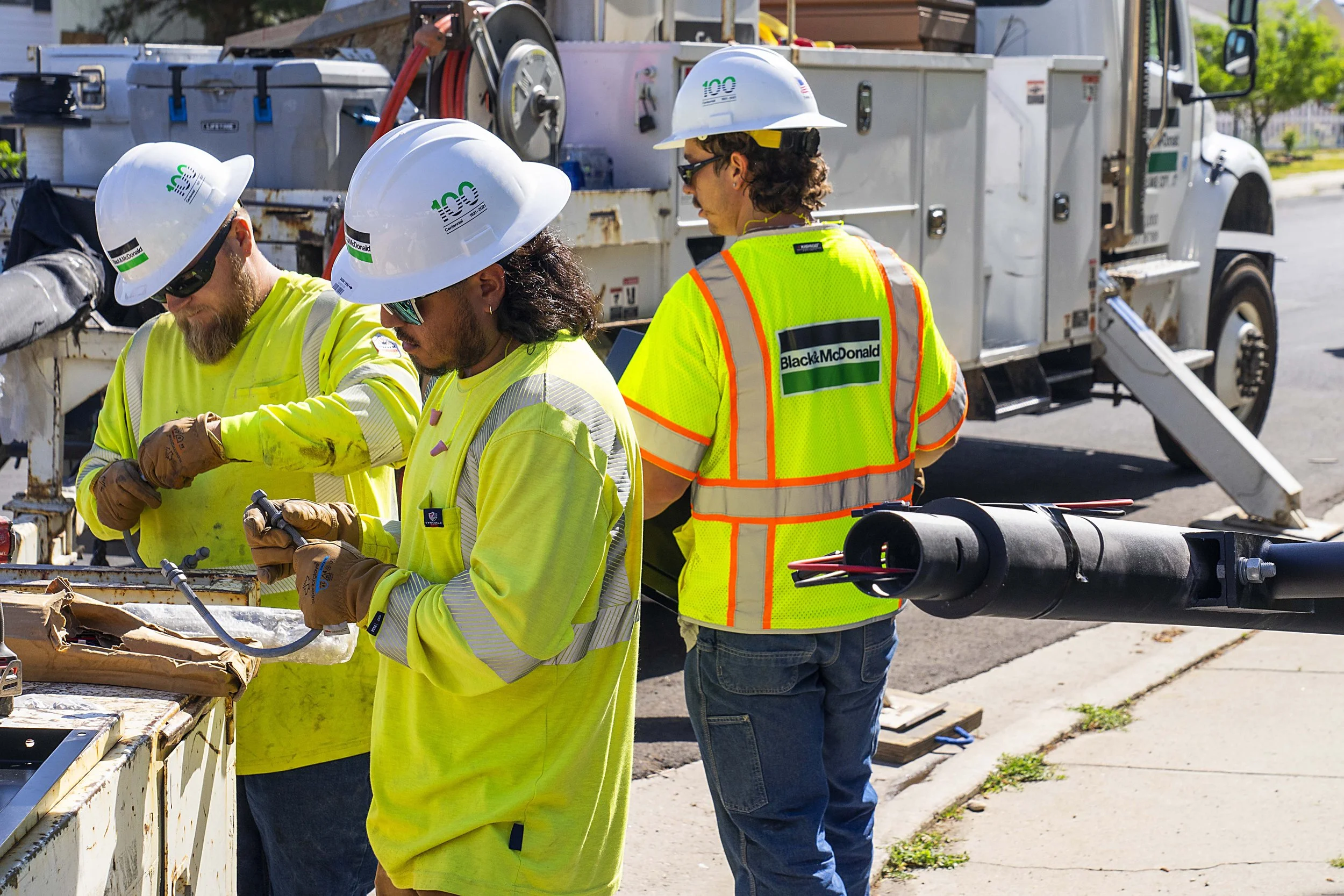 Three construction workers wearing yellow safety vests and white hard hats working near a utility truck on the street.