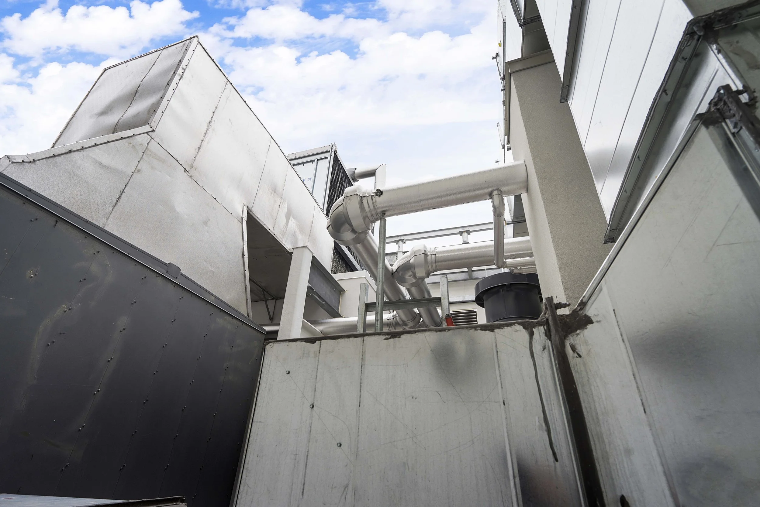 View of industrial rooftop with white and metallic ductwork, pipes, and equipment against a partly cloudy sky.