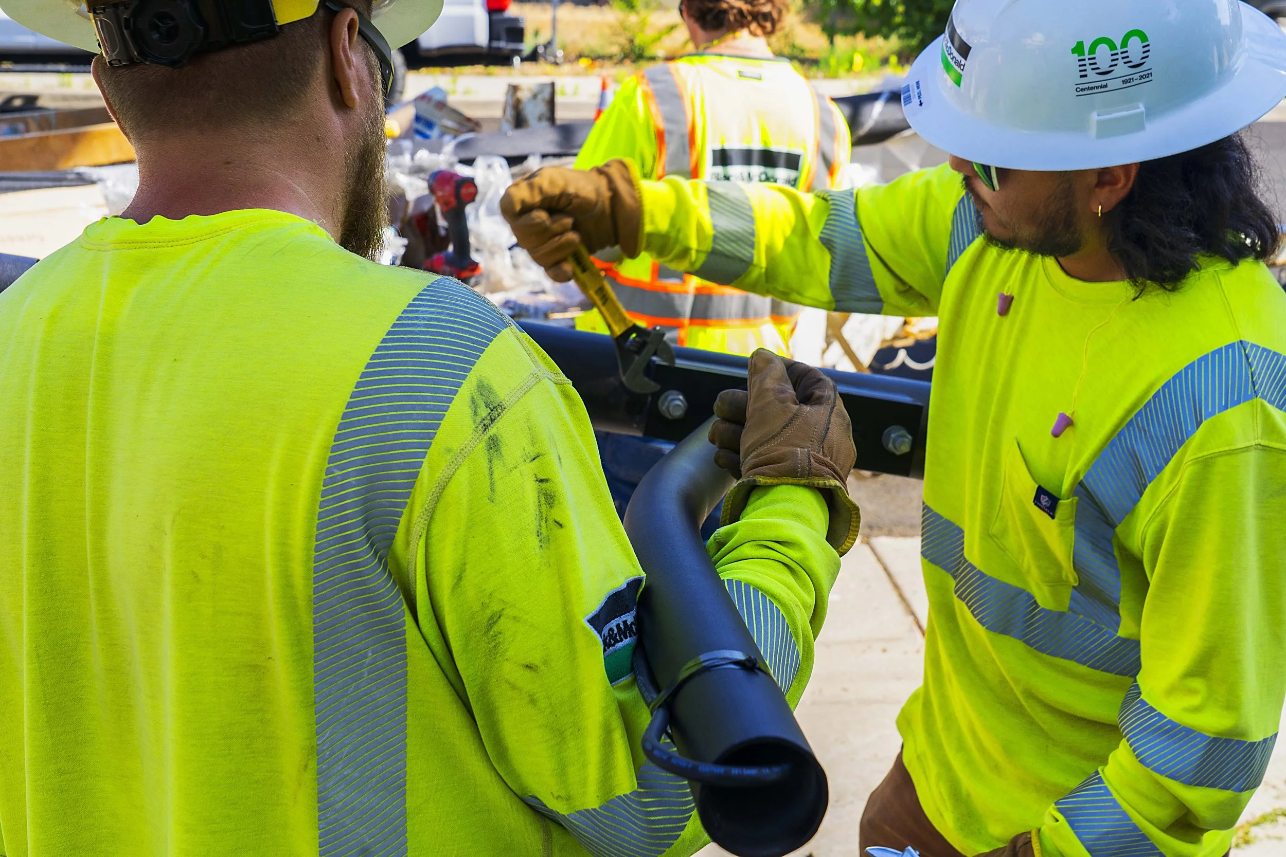 Two construction workers in bright yellow safety vests and helmets working together; one is holding a tool and the other is using a wrench, working on a construction site with various tools and materials in the background.