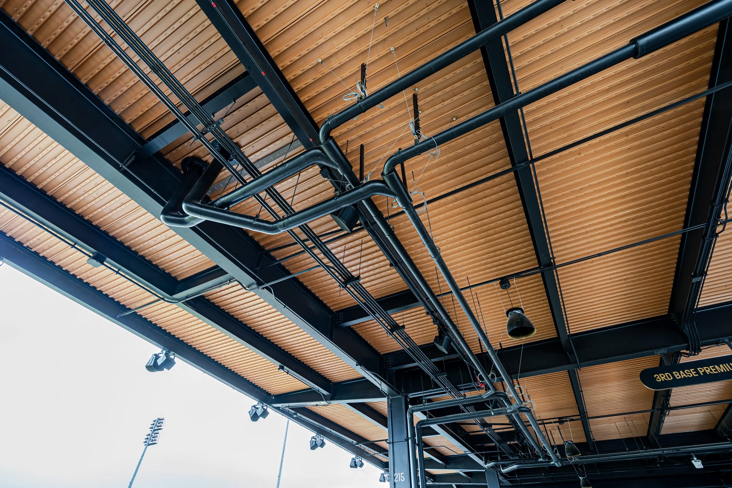 Ceiling of a stadium concourse with black structural beams and orange wood paneling, featuring black pipes, wires, and lighting fixtures.