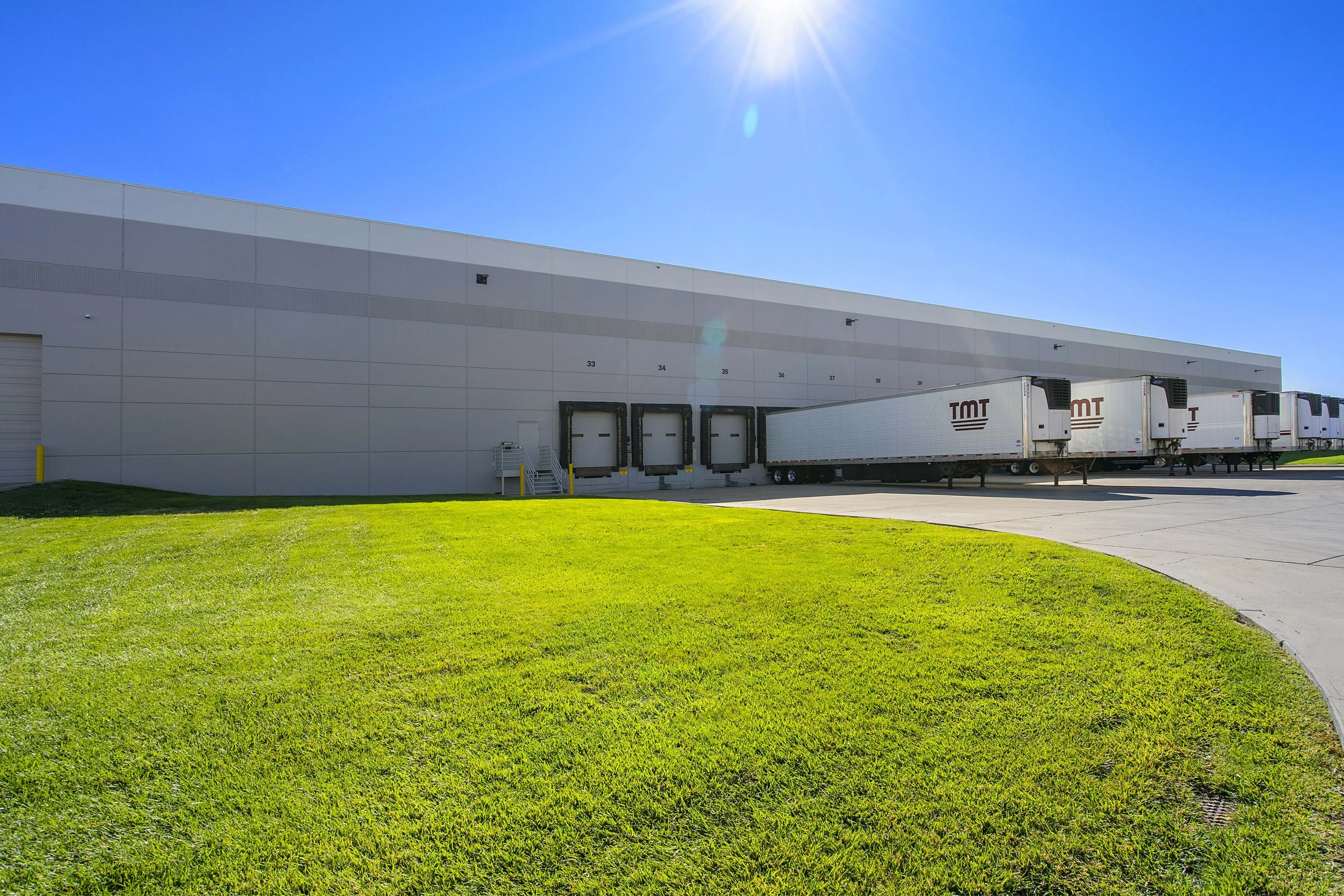 Loading dock with multiple semi-trailer trucks parked in front of a large warehouse under a bright blue sky.