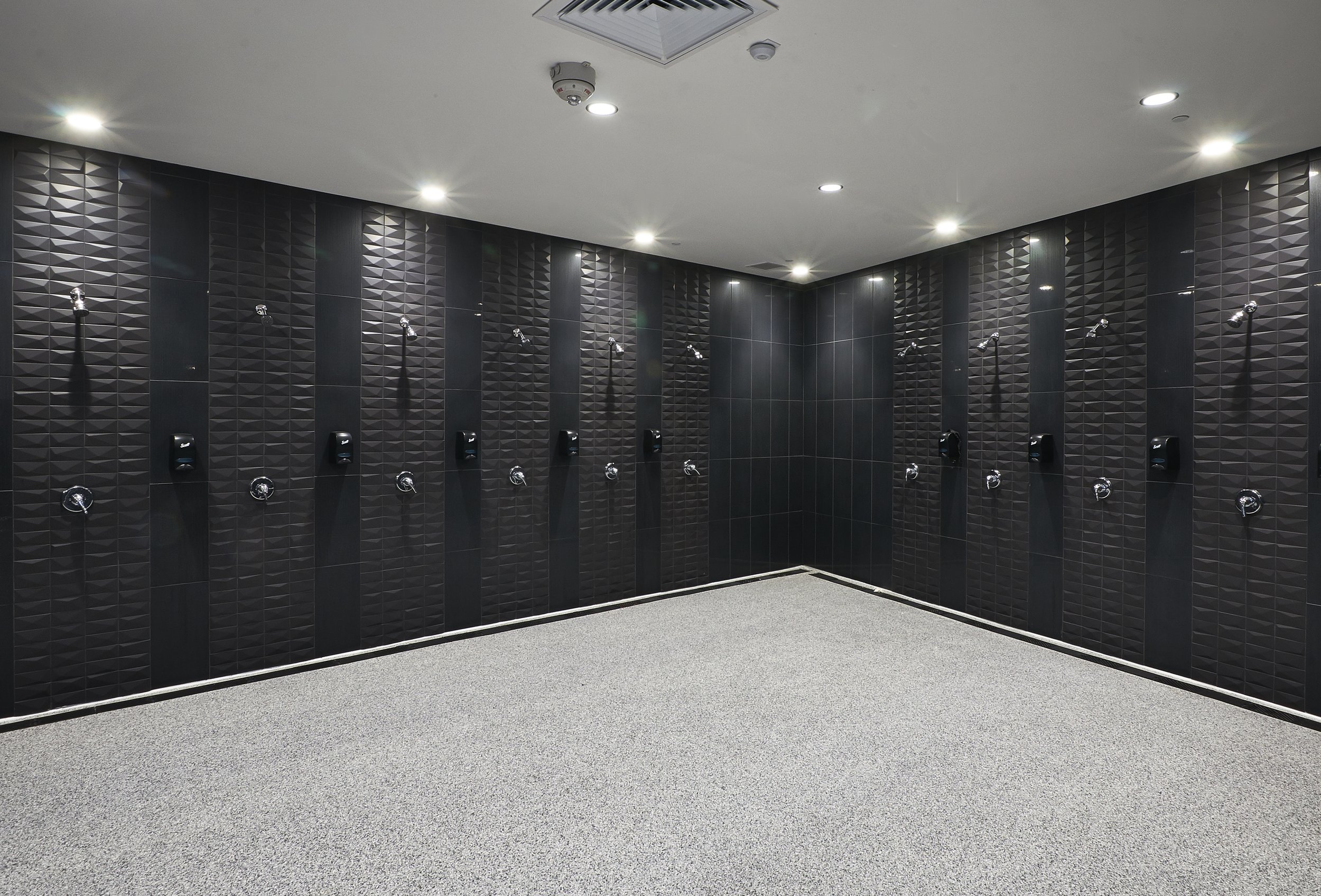 A locker room with black tiled walls and multiple shower heads, soap dispensers, and modern lighting.