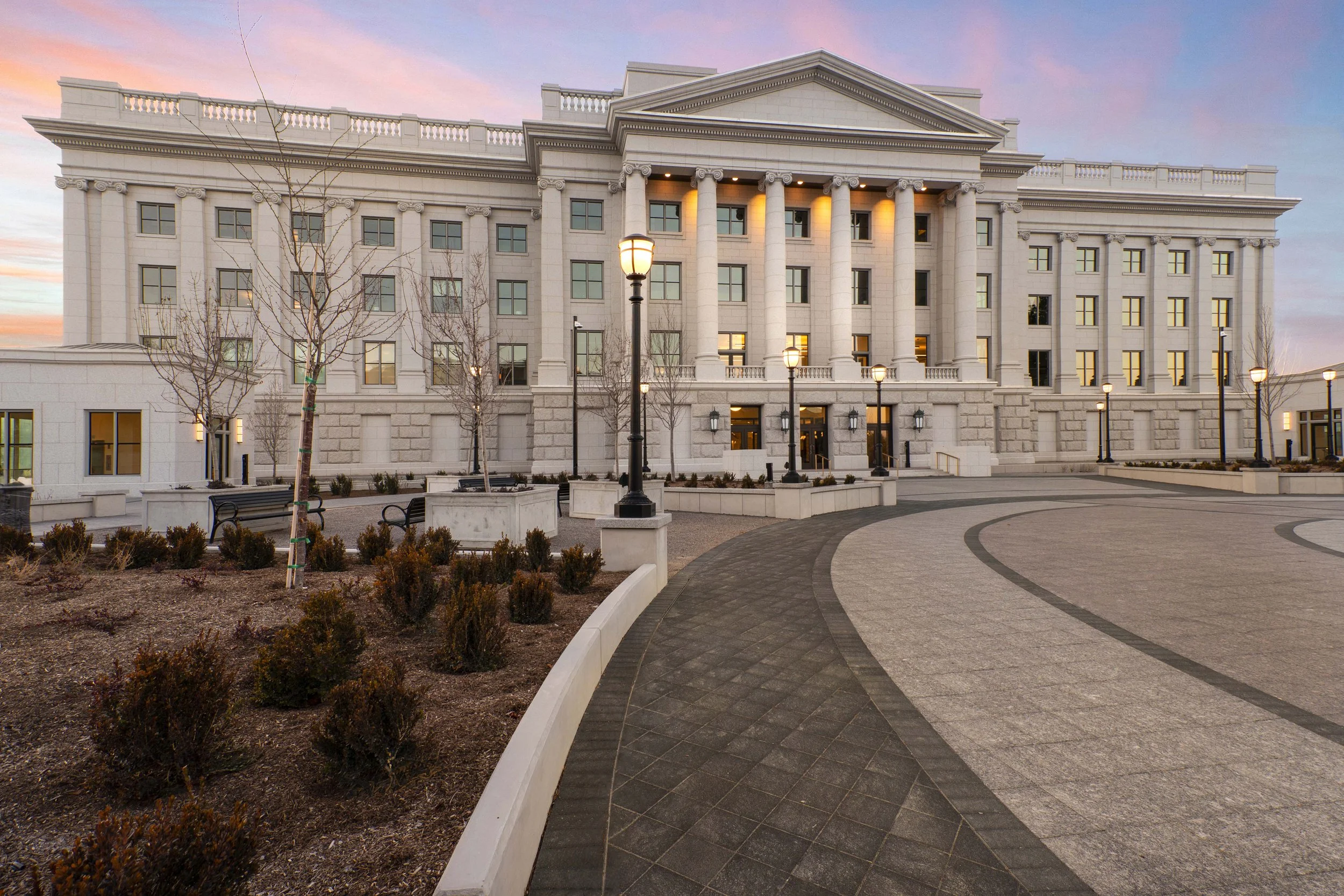 A large federal government building with white stone facade, classical columns, and large windows, set against a pink and blue sky at sunset, with a paved circular driveway, leafless trees, street lamps, and landscaped bushes in the foreground.