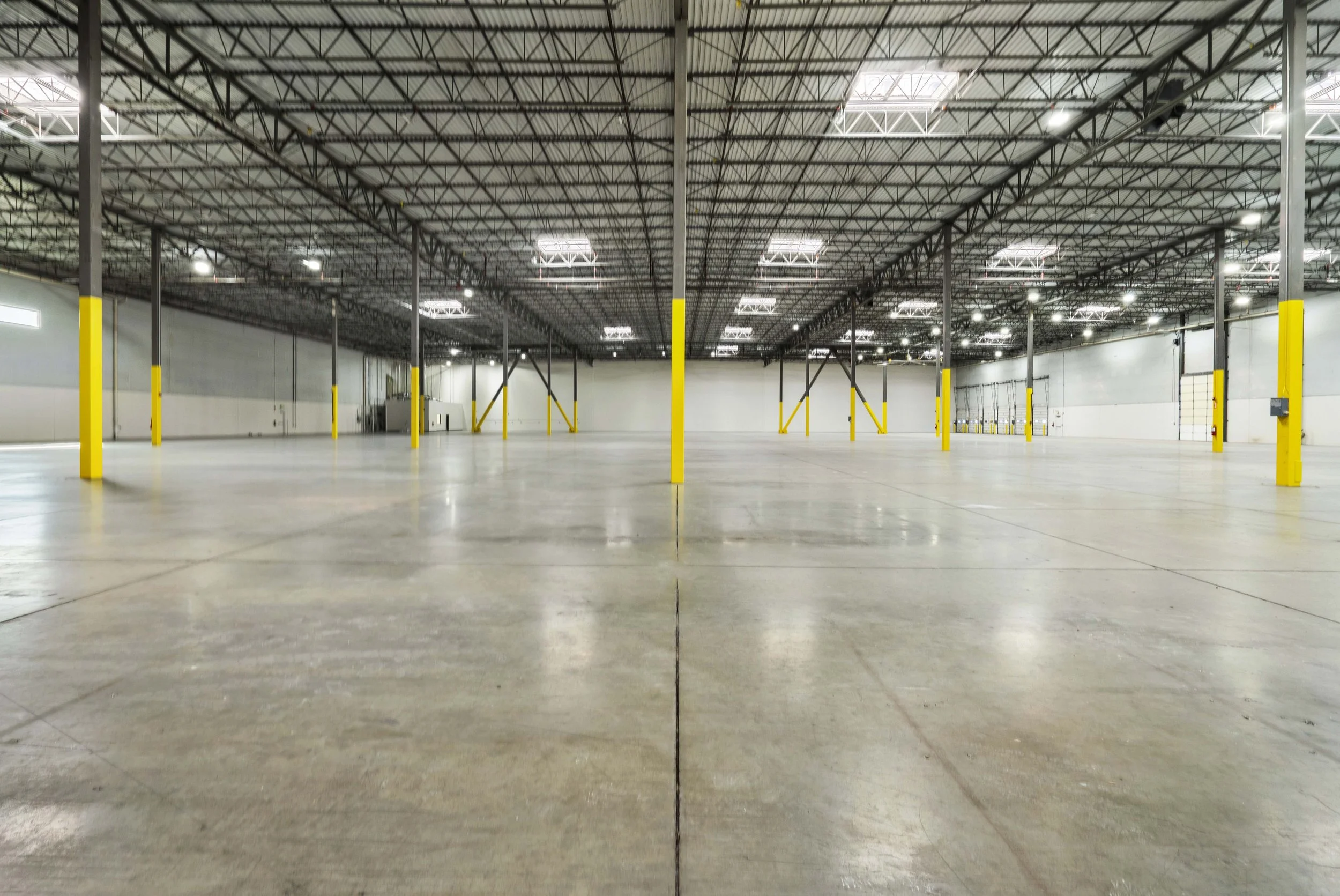 Empty warehouse with concrete floors, tall ceiling with metal beams, and yellow safety posts.