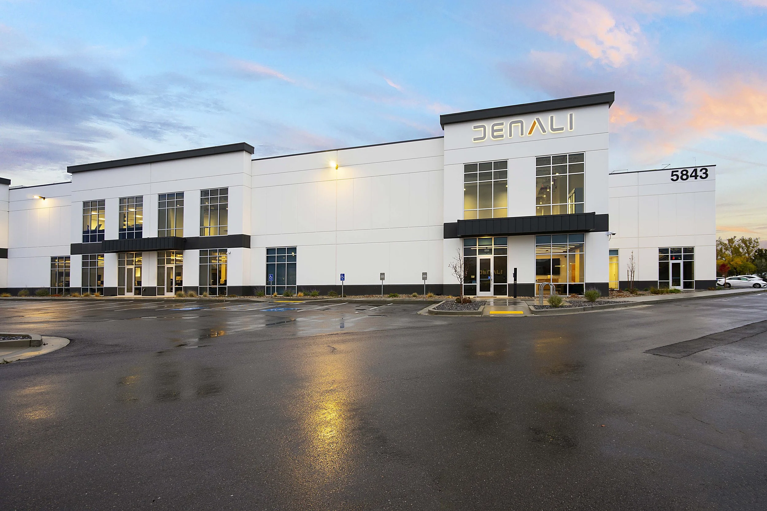 Modern commercial building with a white exterior and black accents, featuring large windows and the sign 'DENALI' on the front, located at 5843. The parking lot in front is wet, with some parked cars and landscaped areas.