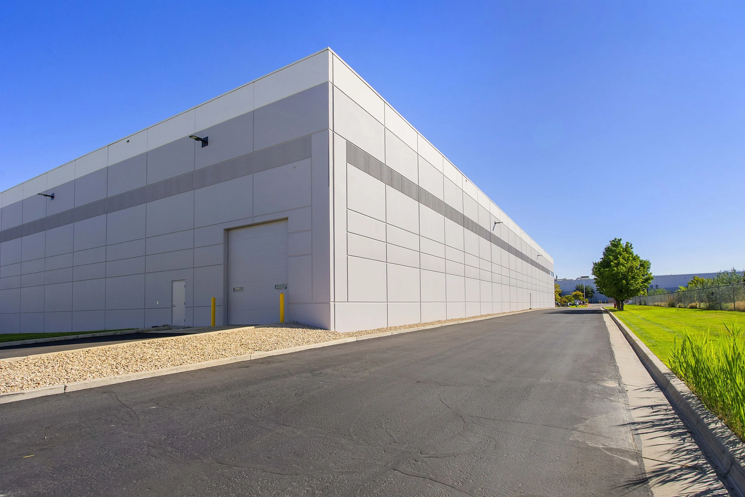Empty commercial warehouse with white walls, large garage door, and a small side door. Paved driveway and green grass with trees under a clear blue sky.