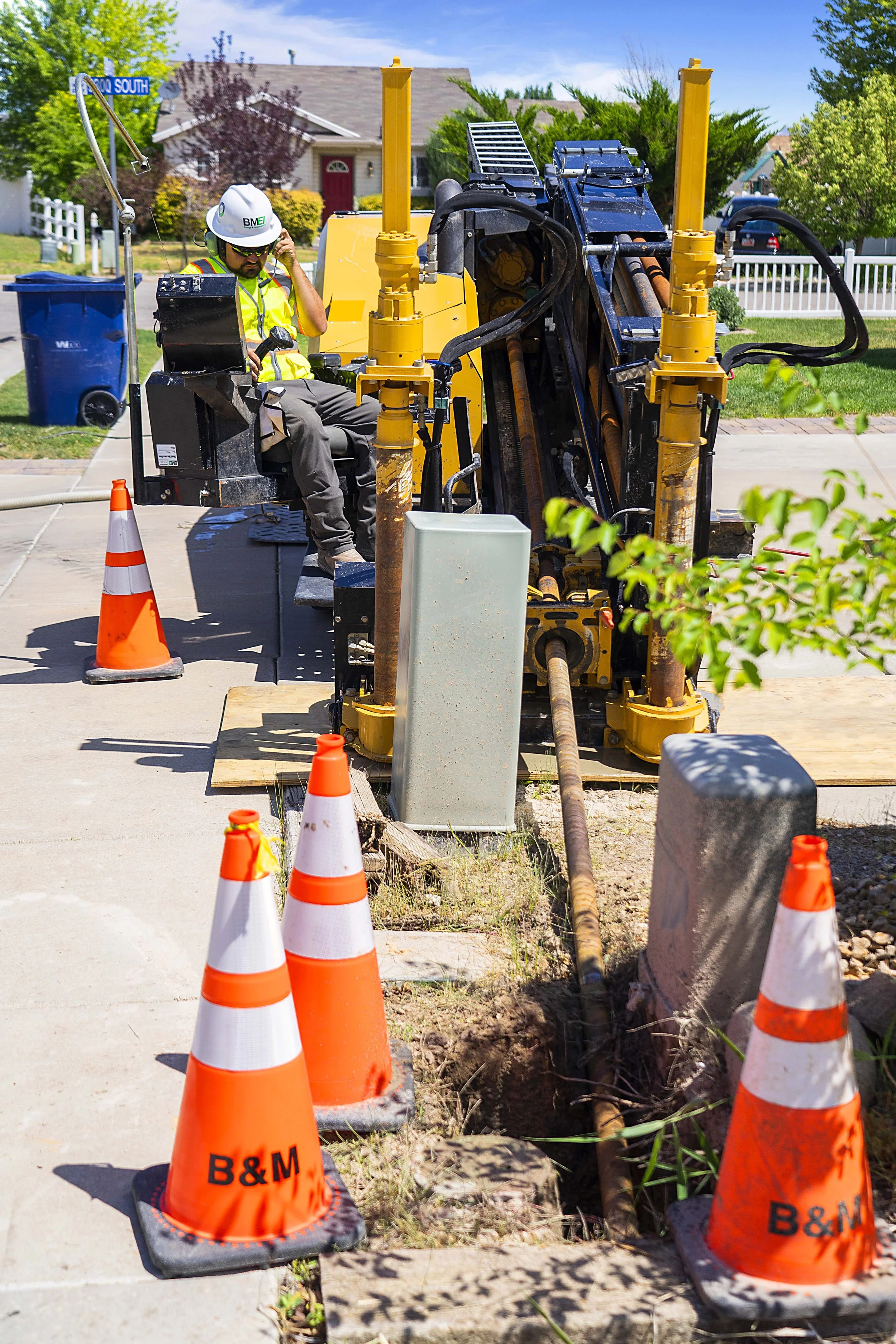 A construction worker in a safety helmet and vest operating a large drilling machine on a residential sidewalk, surrounded by orange safety cones.