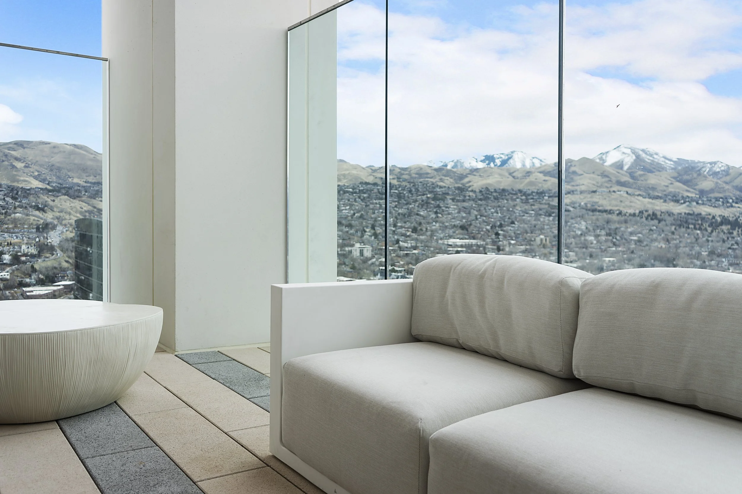 Modern living room with a beige couch and a round coffee table, large floor-to-ceiling windows showing mountains and a partly cloudy sky.