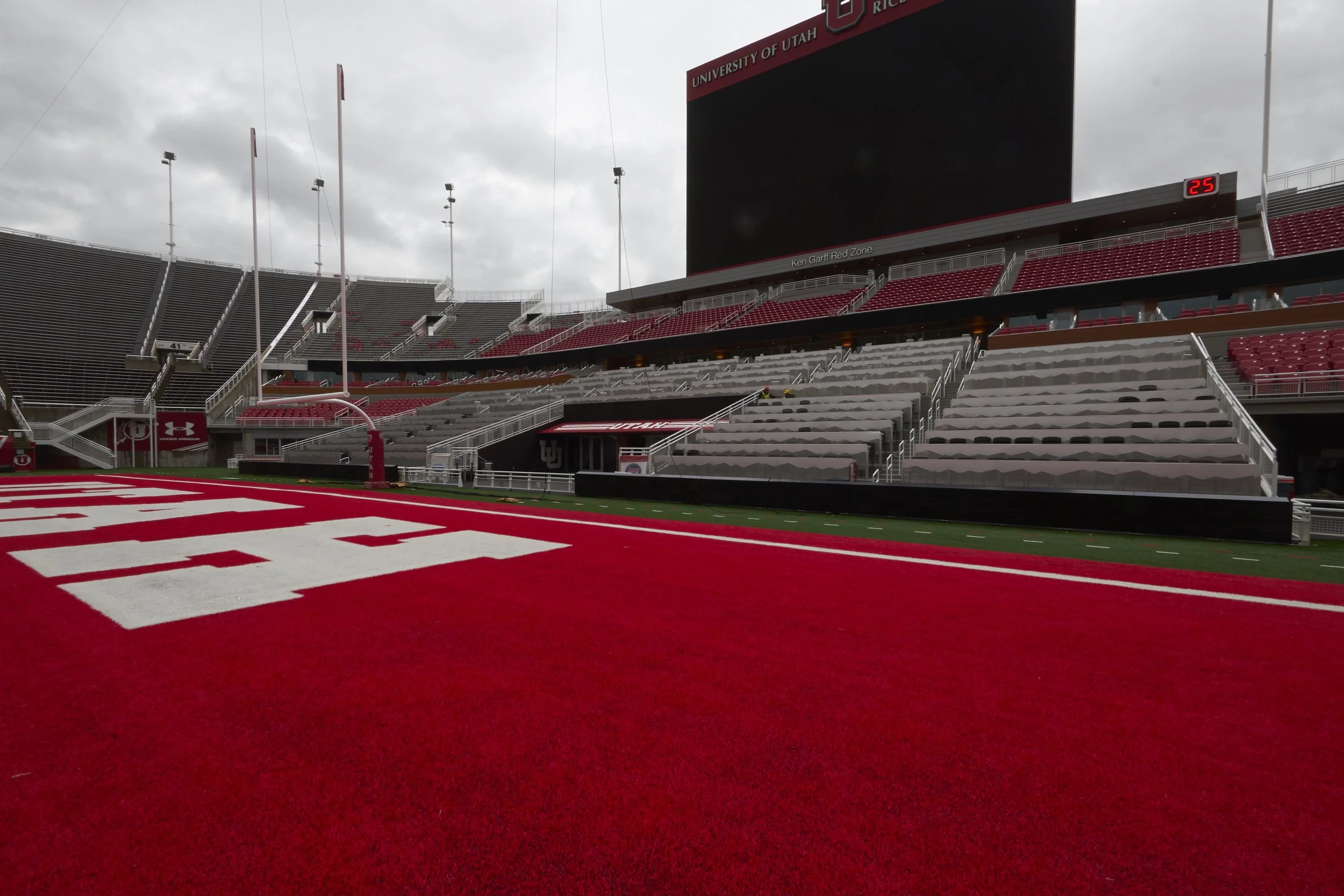 Empty football stadium with red field, white markings, and empty bleachers at University of Utah with overcast sky.