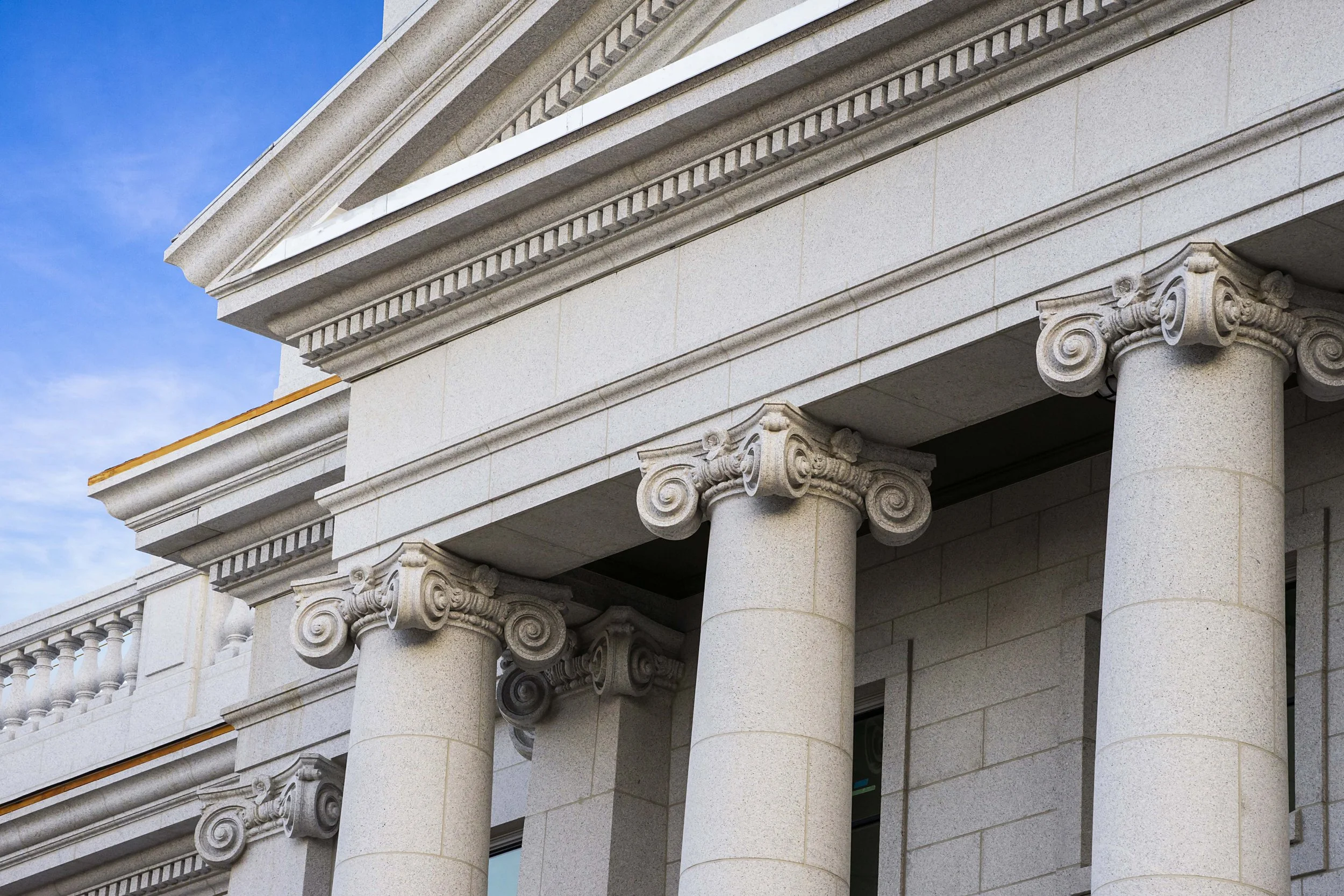 Close-up of a building's classical architecture featuring stone columns with Corinthian capitals and detailed moldings, against a blue sky background.