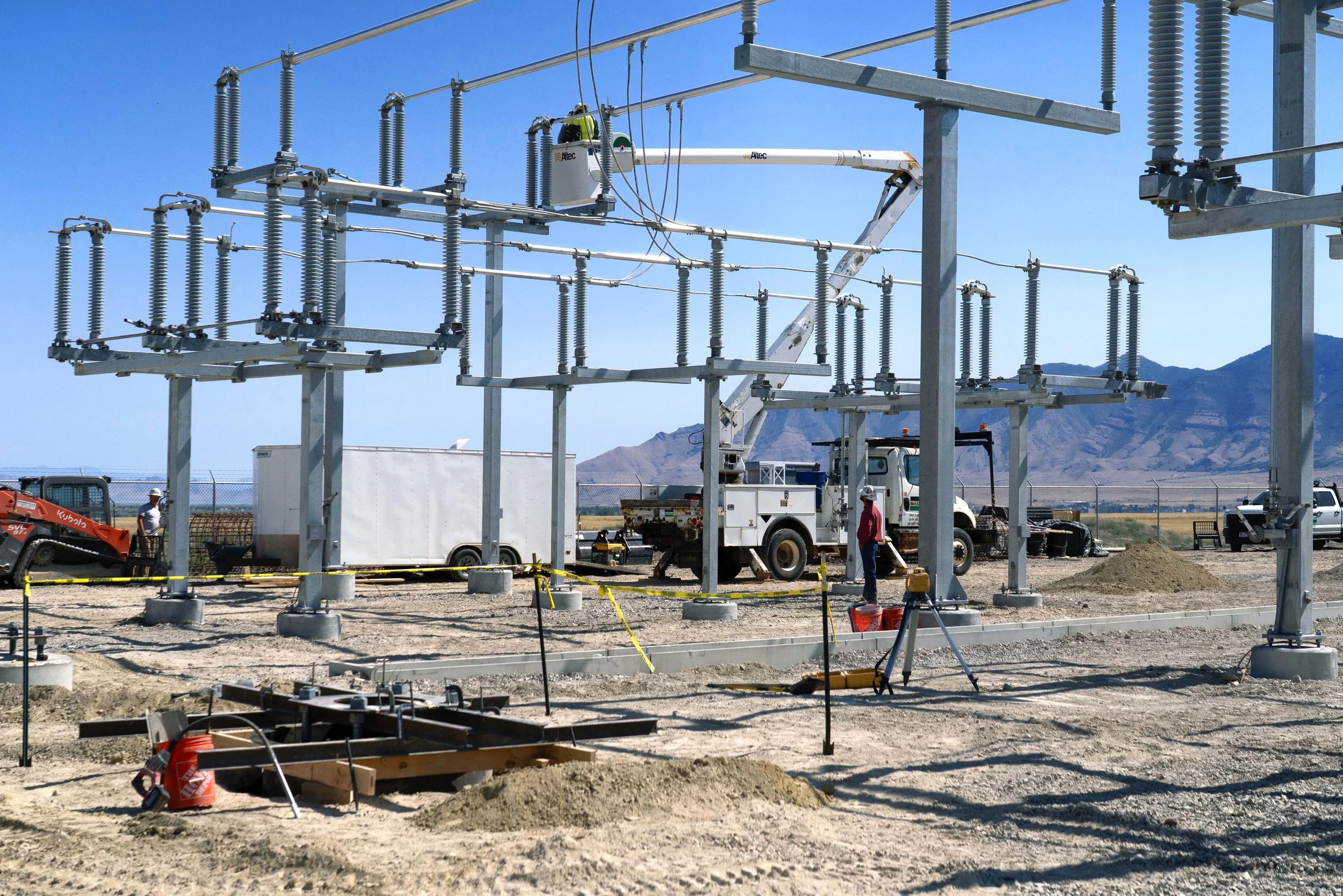 Construction workers installing electrical power lines at an outdoor substation, with mountains visible in the background.