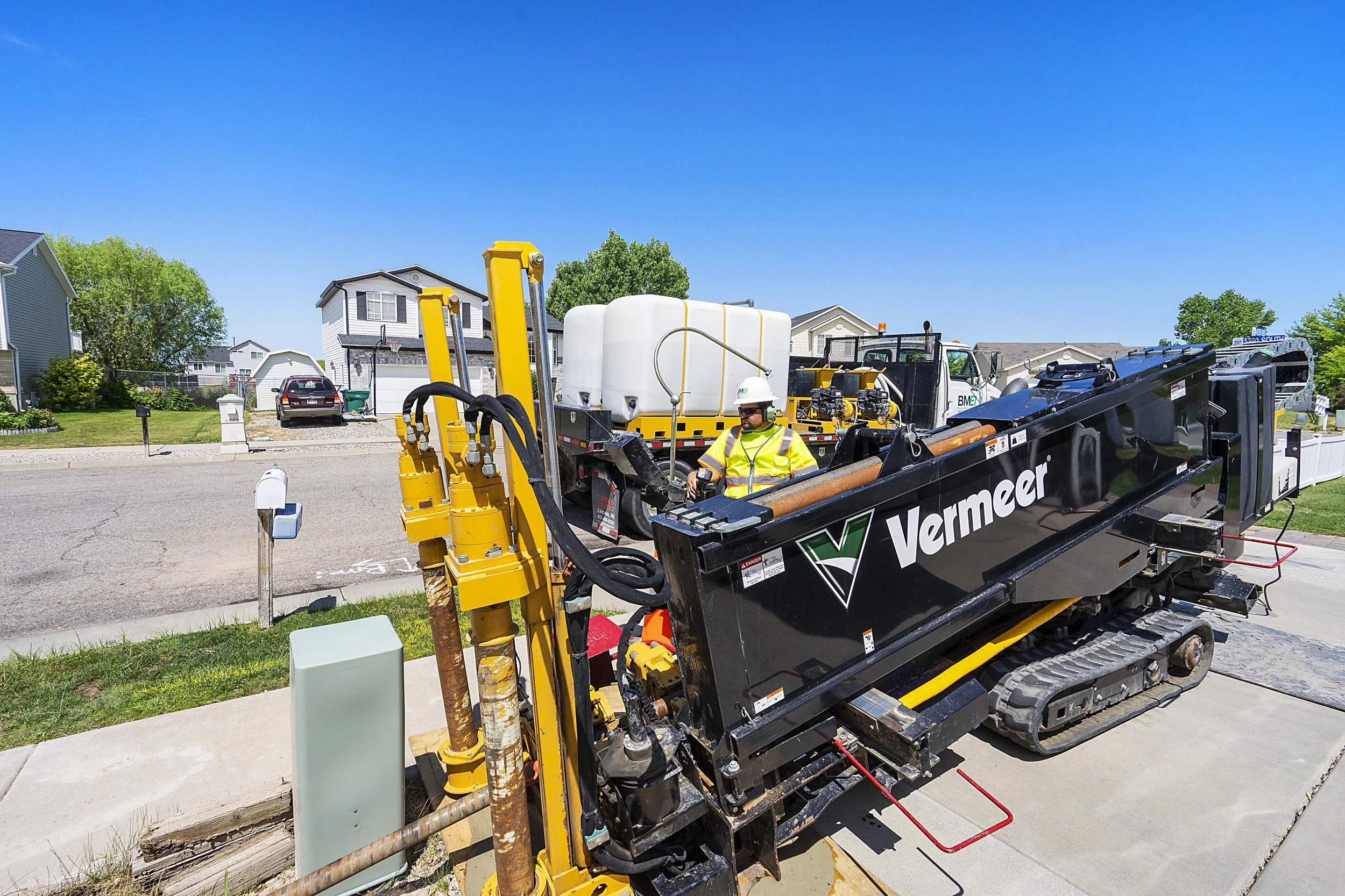 Construction worker in safety gear operating a Vermeer horizontal drilling machine on a residential street with houses and blue sky in the background.