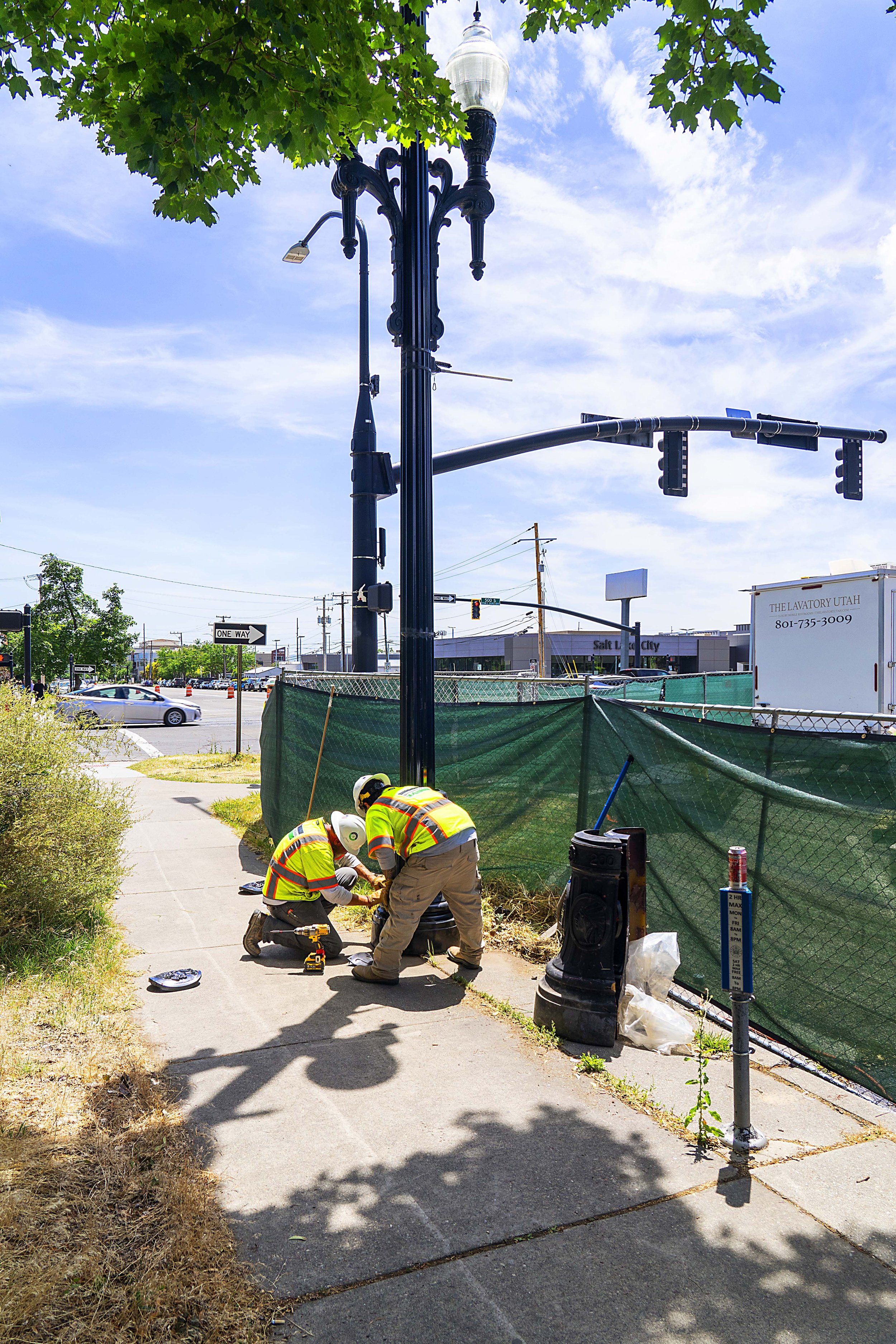 Two workers in safety vests and helmets repairing or installing a streetlamp on a city sidewalk during daytime.