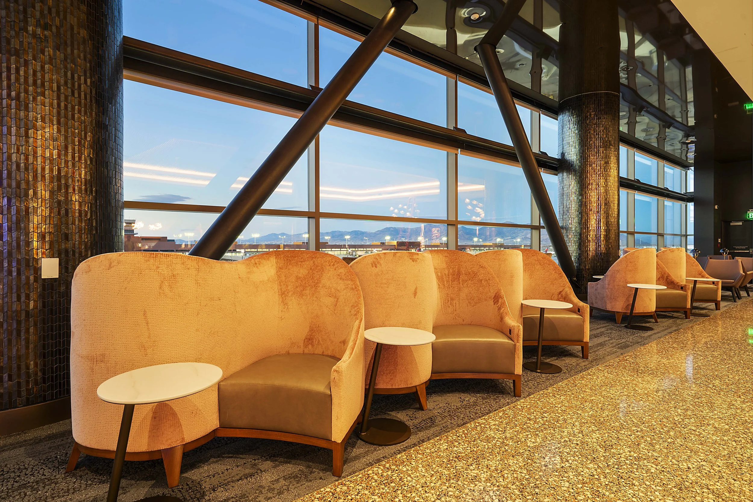 Row of peach-colored armchairs with small side tables inside a modern airport lounge, large windows showing the evening sky with mountains in the distance.