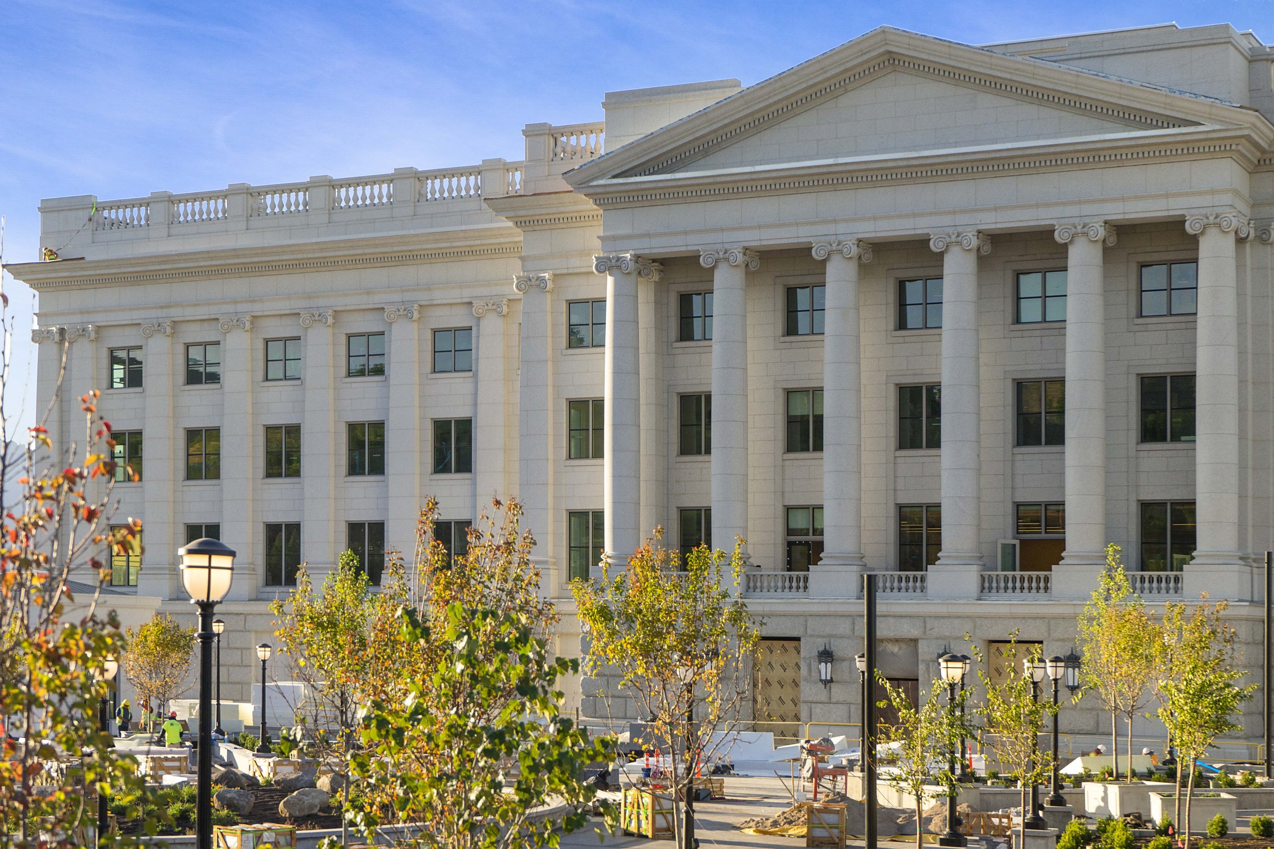 A large neoclassical style white building with tall columns, multiple windows, and a decorative balustrade on top. In front, there is a landscaped area with young trees, lampposts, and ongoing construction.